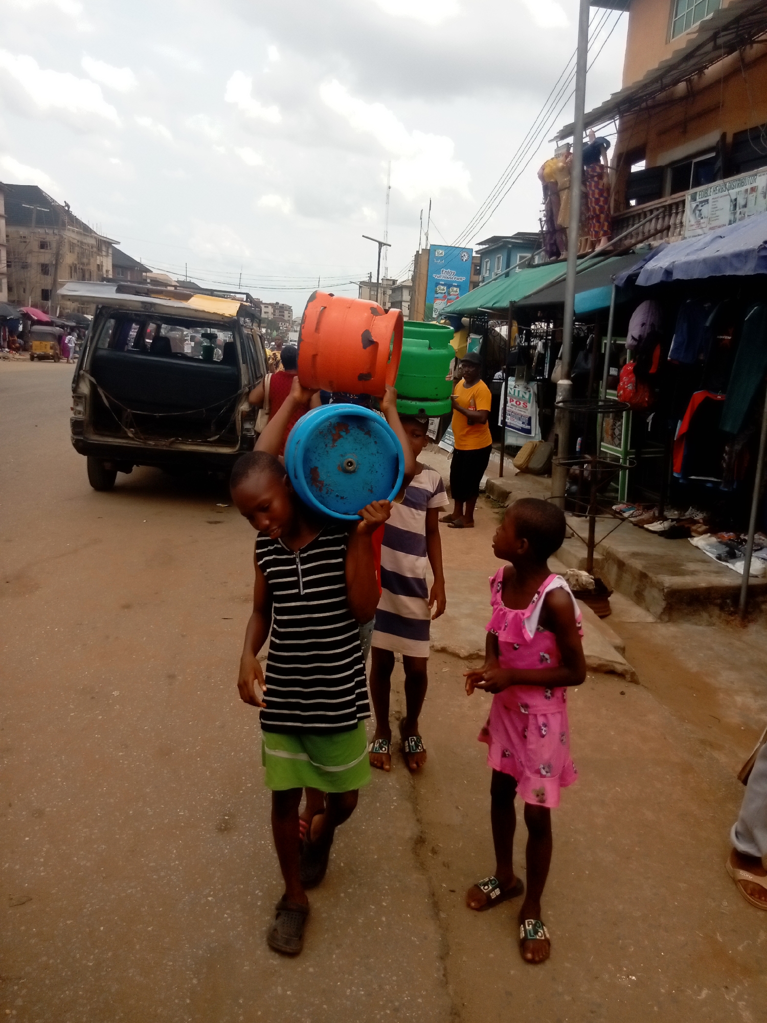 Three children walk on a street in Nkpor, Nigeria. The first child, a boy, carries a blue bucket on his left shoulder and an orange bucket on his head. He is wearing a striped shirt and green shorts. A second child, also a boy, carries a green bucket on his head. He is wearing a striped shirt and no shorts. The third child, a girl, stands near the other two. She is wearing a pink dress. In the background there is a dark-colored sport utility vehicle. Behind the children are shops selling clothes and other goods. There are people visible in the background as well.