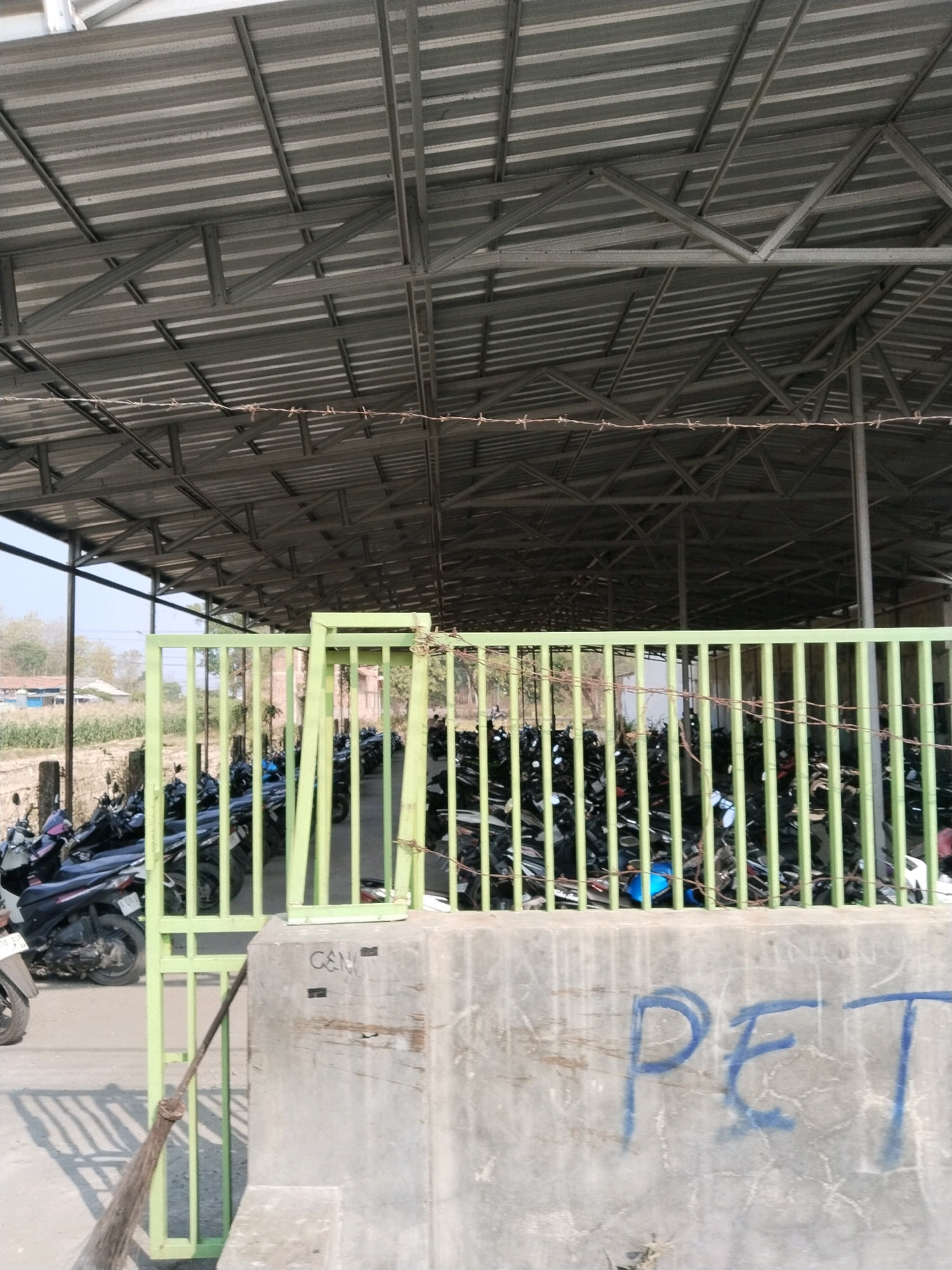 The image depicts a covered parking area for motorcycles in Pakulaut, Indonesia. The structure features a corrugated metal roof supported by a metal frame. A green metal fence encloses the parking area, where numerous motorcycles are parked. The fence is partially obscured by barbed wire. In the foreground, a concrete barrier with graffiti reading "PET" is visible. A broom leans against the fence next to the barrier. The environment appears to be outdoors, with some vegetation and buildings visible in the background. The time of day is likely daytime, indicated by the sunlight. There are no people clearly visible in the image.