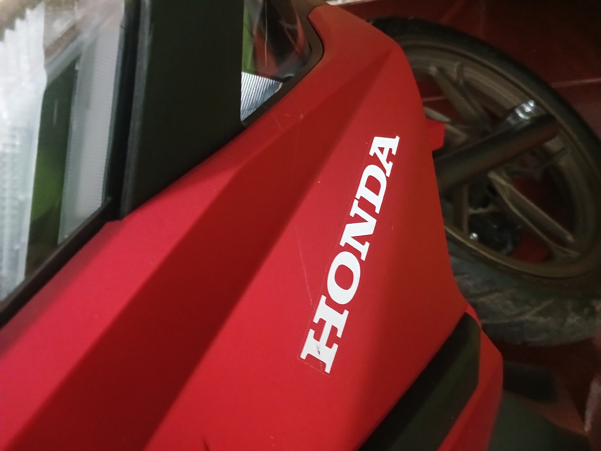 The image is a close-up, eye-level shot of the side of a red Honda motorcycle. The word "HONDA" is visible in white lettering on the motorcycle's fairing. A portion of the front wheel and tire, along with part of the suspension, are also visible. The background is dark and out of focus, but appears to be indoors. The scene is consistent with a photograph taken in Randuagung, Indonesia, where Honda motorcycles are commonly used.