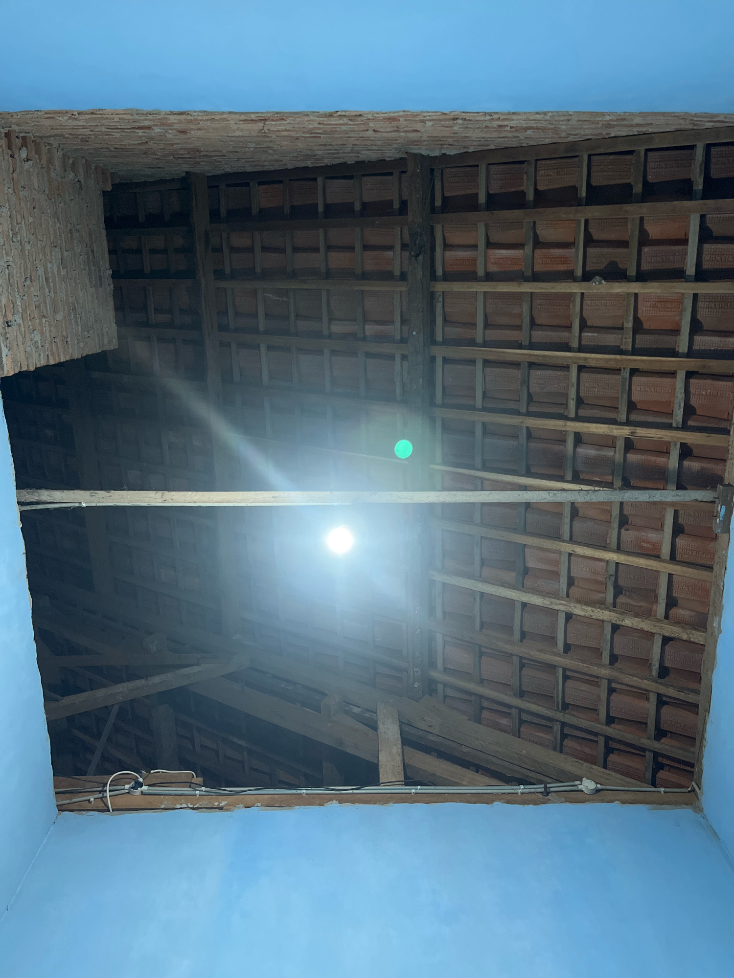 The image depicts the interior of a partially constructed or unfinished building, likely in Kebumen, Indonesia, given the context. The ceiling, which is the primary focus, is composed of exposed wooden beams supporting rows of terracotta roof tiles. A single bright light fixture hangs from one of the beams, illuminating the space. The walls are painted a light blue. The scene suggests an ongoing construction or renovation project. There are no people or animals visible in the image.