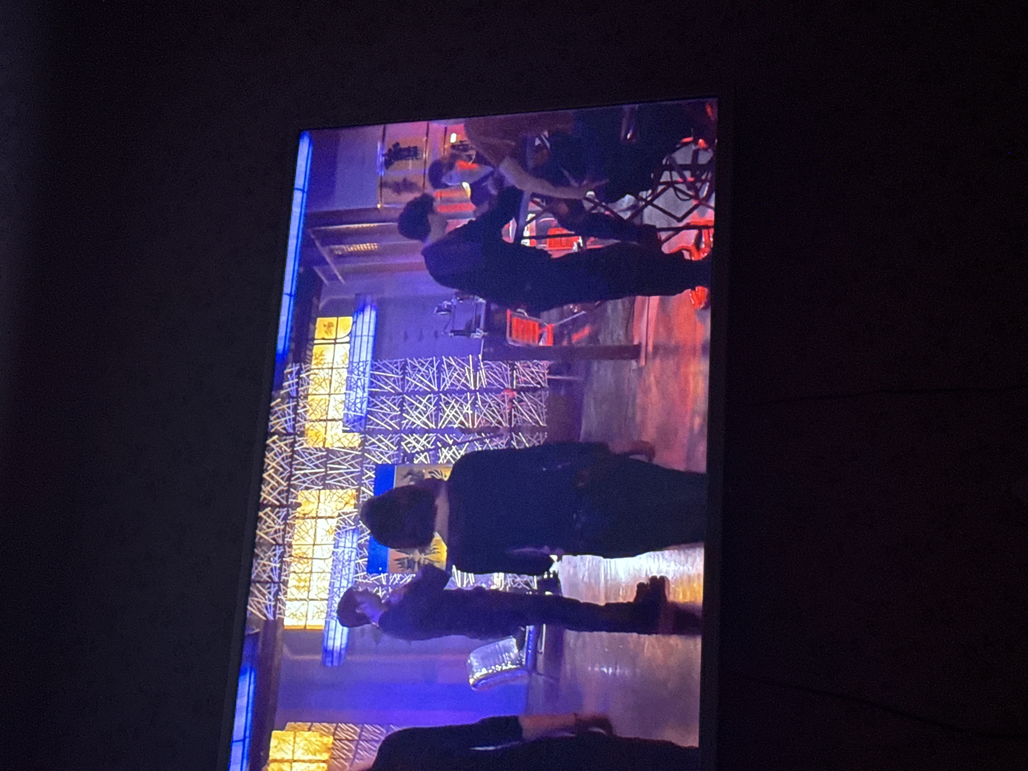 The image shows a television screen displaying a scene with multiple people. The subjects are dressed in dark clothing. In the background, there is an illuminated structure featuring blue and gold colors. There are also metal scaffolding elements in the scene. No information is provided that identifies the location as Mytishchi, Russia.