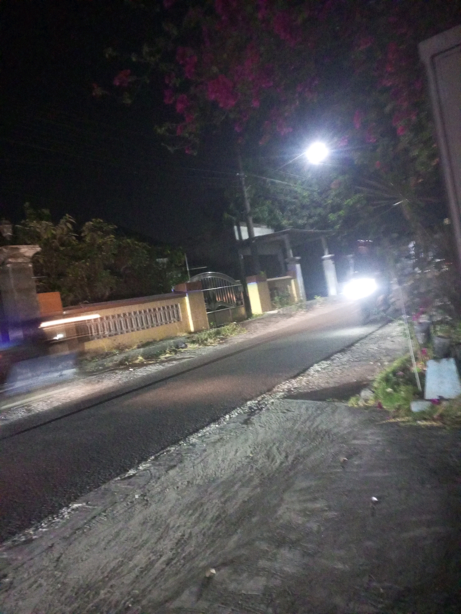 The image captures a street scene at night in Ngasem, Indonesia. The foreground shows a paved road with a rough texture on the right shoulder, where scattered debris is visible. To the left, the road is smoother and reflects light from above. In the midground, a low yellow wall with decorative railings runs parallel to the road, bordered by a grassy verge. Beyond the wall, trees and foliage are dimly lit. A streetlamp illuminates the scene from the upper right quadrant. A white vehicle with bright headlights is moving away from the viewer on the road. The background is dark, with hints of structures and more foliage, and pink flowers are visible in the upper portion of the image. The overall impression is of a quiet, dimly lit residential street at night.
