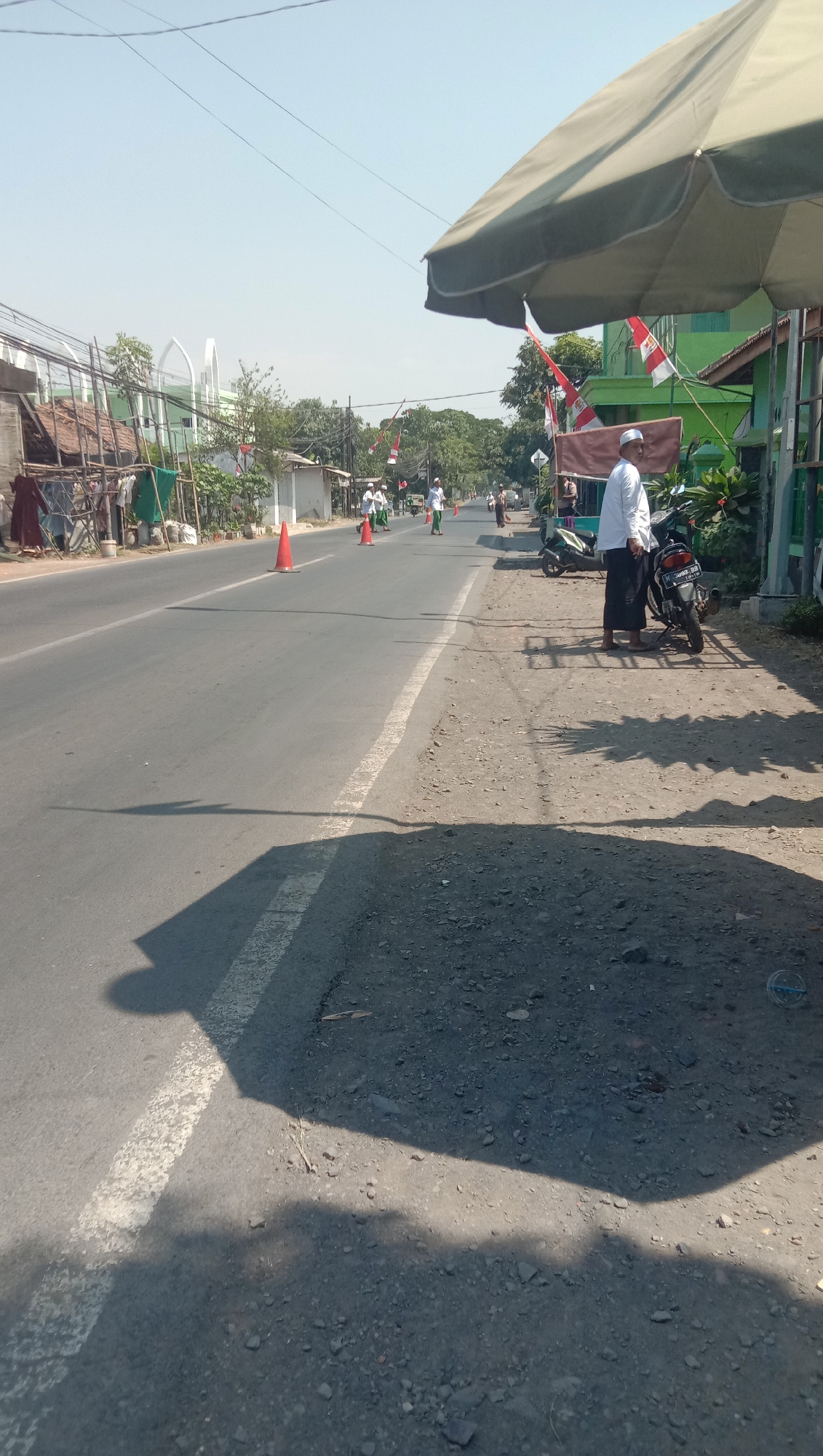 In Karang Anyar, Indonesia, a man wearing a white shirt and sarong, and a peci, stands by a parked motorcycle on a dusty shoulder next to a paved road. The road is lined with flags, including Indonesian flags, and traffic cones are placed along the roadway. Several other men, similarly dressed, walk along the road in the distance. The scene is under a large, light-colored umbrella, casting a shadow on the ground. Buildings, including a green structure with a red roof and some wooden scaffolding, are visible on the side of the road. The sky is clear and blue, indicating a sunny day.