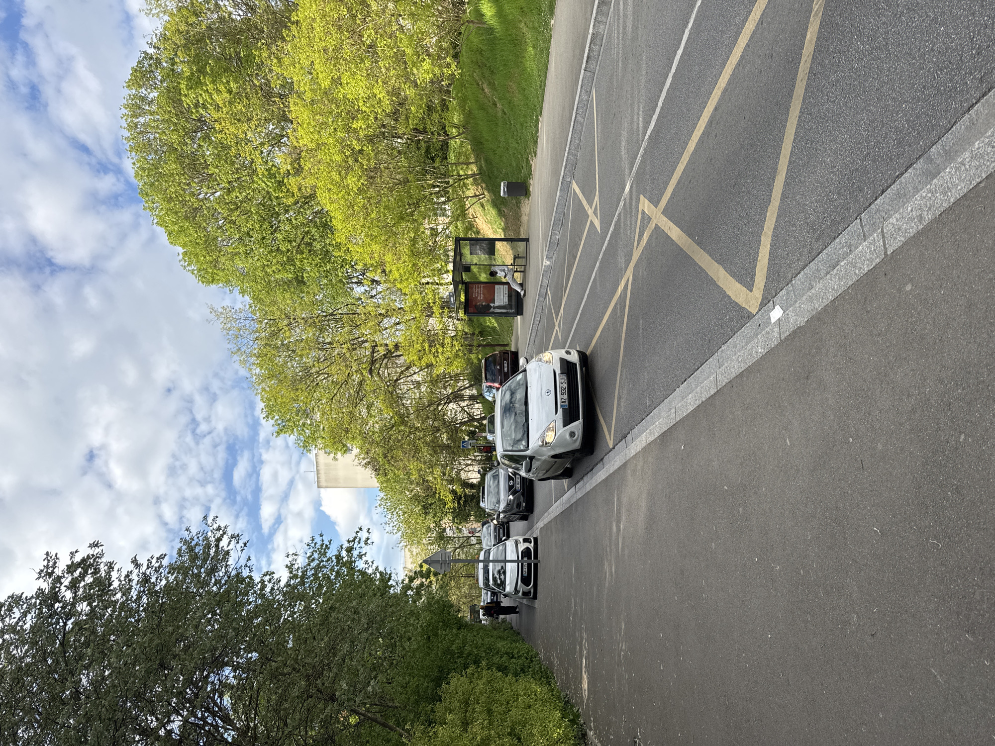 The image depicts a street scene in Les Ulis, France. Cars are parked along the right side of the road, facing forward. A bus stop is located behind the cars, with a person sitting inside. The road has yellow crosshatch markings, indicating a restricted area. Trees and greenery line the left side of the road. The sky is visible with scattered clouds. A building is located in the background.