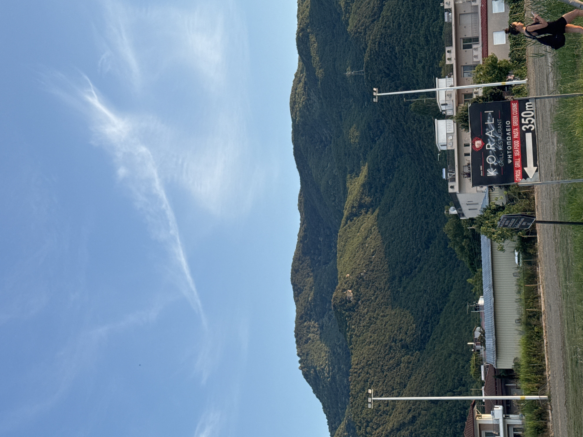 The image depicts a scenic view of the city of Stavros, Greece, with a majestic mountain range serving as a backdrop. The sky is a clear blue, adorned with wispy white clouds, suggesting fair weather. In the foreground, a street lamp stands tall, illuminating the path. A sign for KORALI GRILL-RESTAURANT, which specializes in Gyros, Grill, Sea Food, Pasta, and Greek Cuisine, indicates that it is 350 meters away. A woman in a black dress walks by, holding a smartphone in her hand. The buildings in the background are typical of Greek architecture, with white walls and tiled roofs. The lush green vegetation surrounding the area adds to the serene ambiance of the scene.
