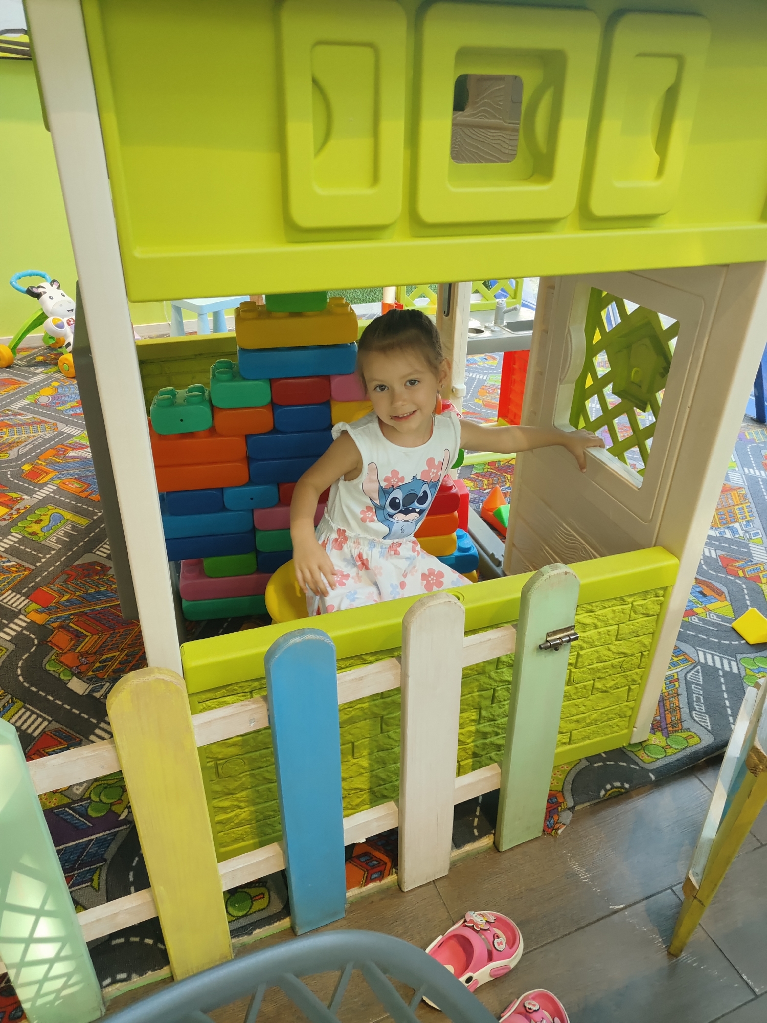 A young girl with brown hair and a white t-shirt with Stitch on it and floral shorts is sitting inside a brightly colored plastic playhouse. She is smiling and looking at the camera. The playhouse is made of yellow and white plastic and has a green fence in front of it. Inside the playhouse are large, colorful plastic blocks. The floor is covered with a colorful rug with a city street pattern. In the background, there are other toys, including a zebra push toy. The scene appears to be indoors, likely in a children's play area, possibly in the city of Chornomorsk, Ukraine, given the context.