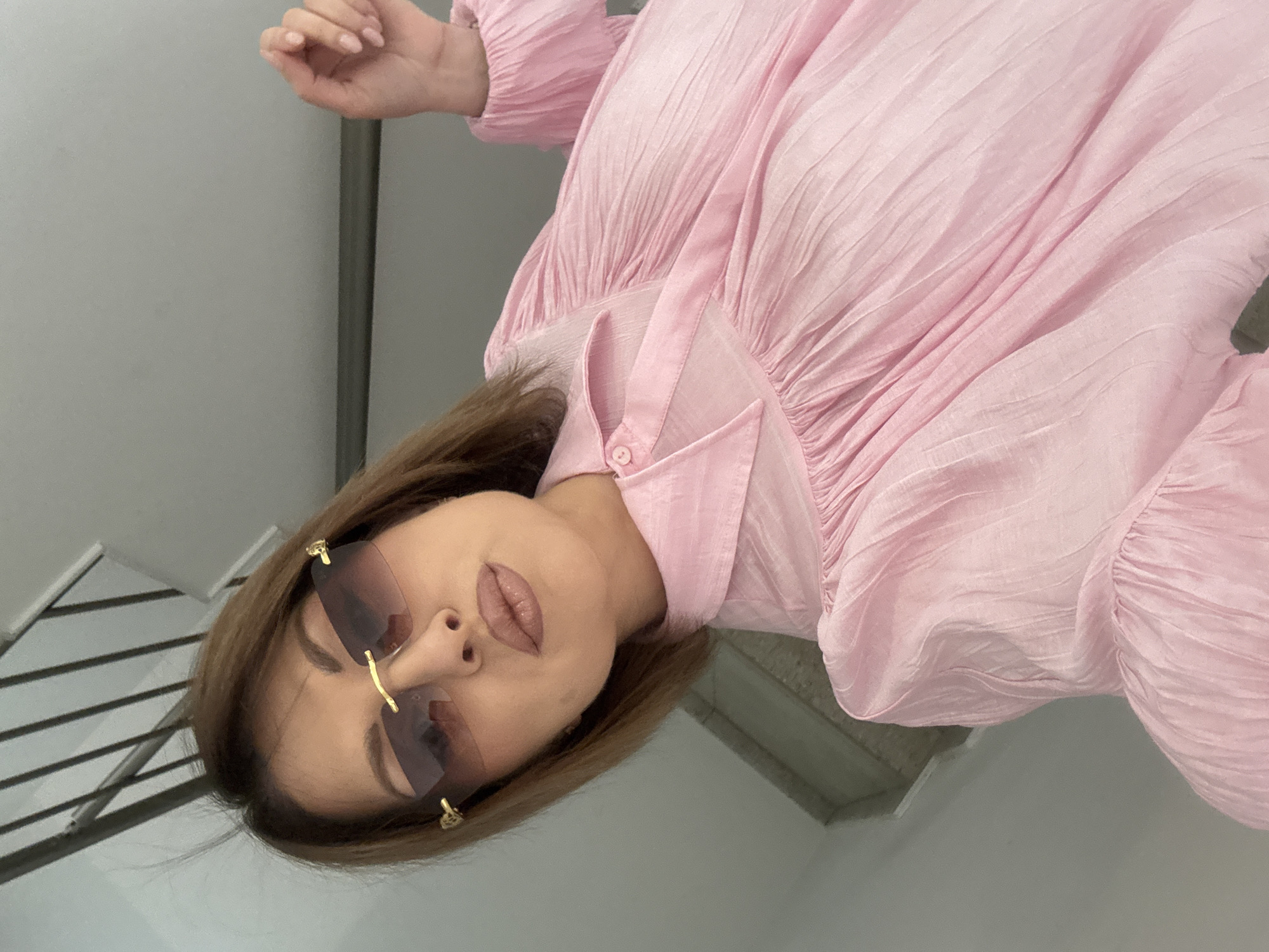 A woman wearing sunglasses and a pink dress is photographed indoors, with her head tilted. The location appears to be a staircase next to a gray wall. No specific details indicate that it is in Sofia, Bulgaria.