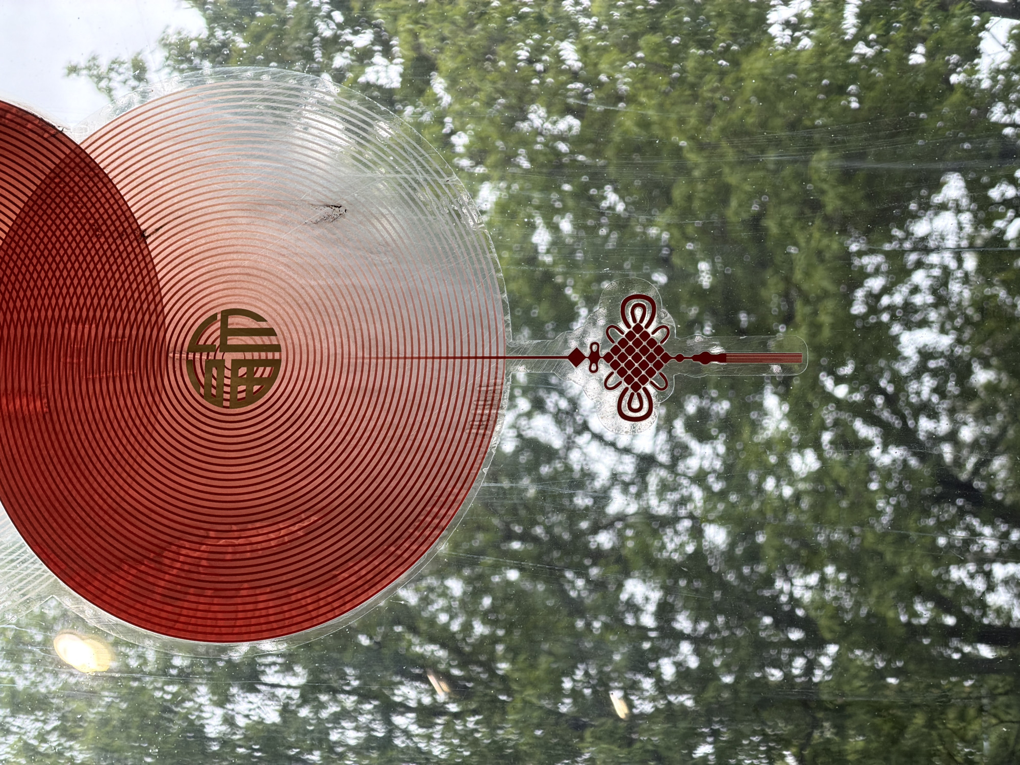 The image depicts an ornate, circular sticker adhered to a window, partially obscuring a view of trees in Saint Petersburg, Russia. The sticker is predominantly red and white, featuring concentric circles radiating from a central golden-colored emblem. To the right, a stylized knot symbol extends from the circular design. The window is slightly dirty, with visible streaks and smudges. Outside, the blurred greenery of trees fills the background, characteristic of Saint Petersburg's urban landscape. The scene appears to be captured indoors, looking out towards the park.