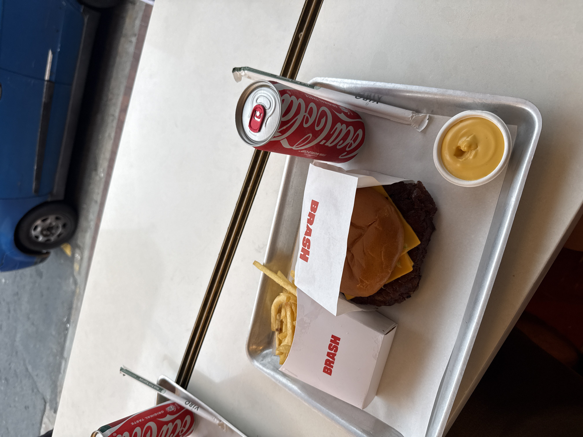 On a metal tray, a Brash burger with cheese and fries, a can of Coca-Cola, and a small container of yellow sauce are placed. The tray is positioned on a white table indoors. Through the window, a blue car and a sidewalk scene are partially visible. The scene is located in Cape Town, South Africa.