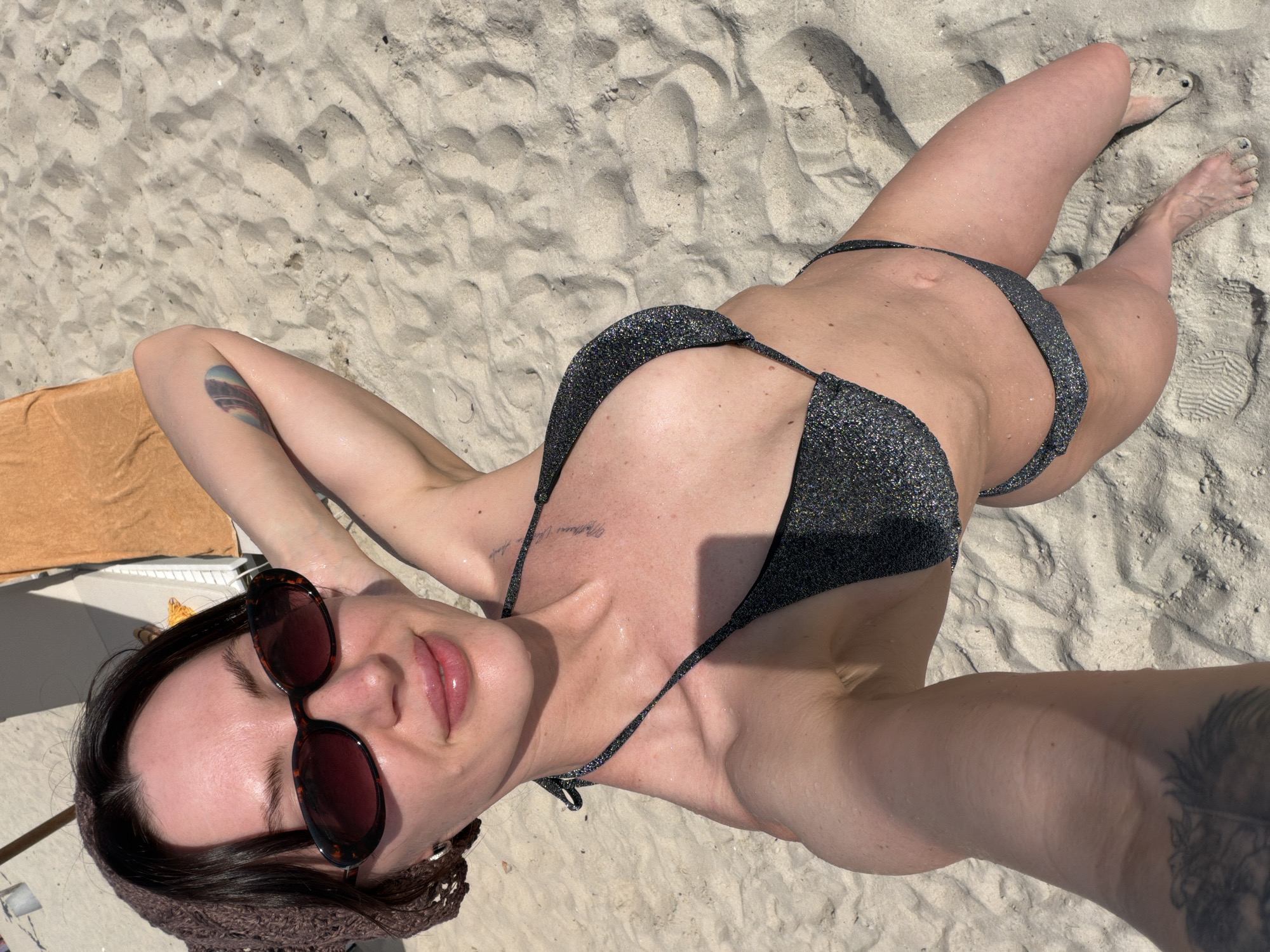 An adult female is depicted from a high-angle perspective, engaged in a self-portrait on a sandy beach. She is wearing a dark, glittery two-piece bikini, consisting of a halter-neck top and string bottoms. Her dark hair is partially covered by a brown, woven headpiece, and she wears oval tortoiseshell sunglasses. A small, silver hoop earring is visible on her left ear. Her lips appear glossy. Visible tattoos include faint script on her upper left chest and a multi-colored abstract design on her inner left upper arm. Another large, dark tattoo is partially visible on her lower right leg. Her skin displays a moist sheen. The immediate environment consists of light-colored sand with numerous footprints and texture. In the background, a white lounge chair draped with an orange towel and a partial beach umbrella structure are discernible. The scene is outdoors, under bright natural light. This beach is situated in Dong Ninh Hoa Ward, Vietnam.