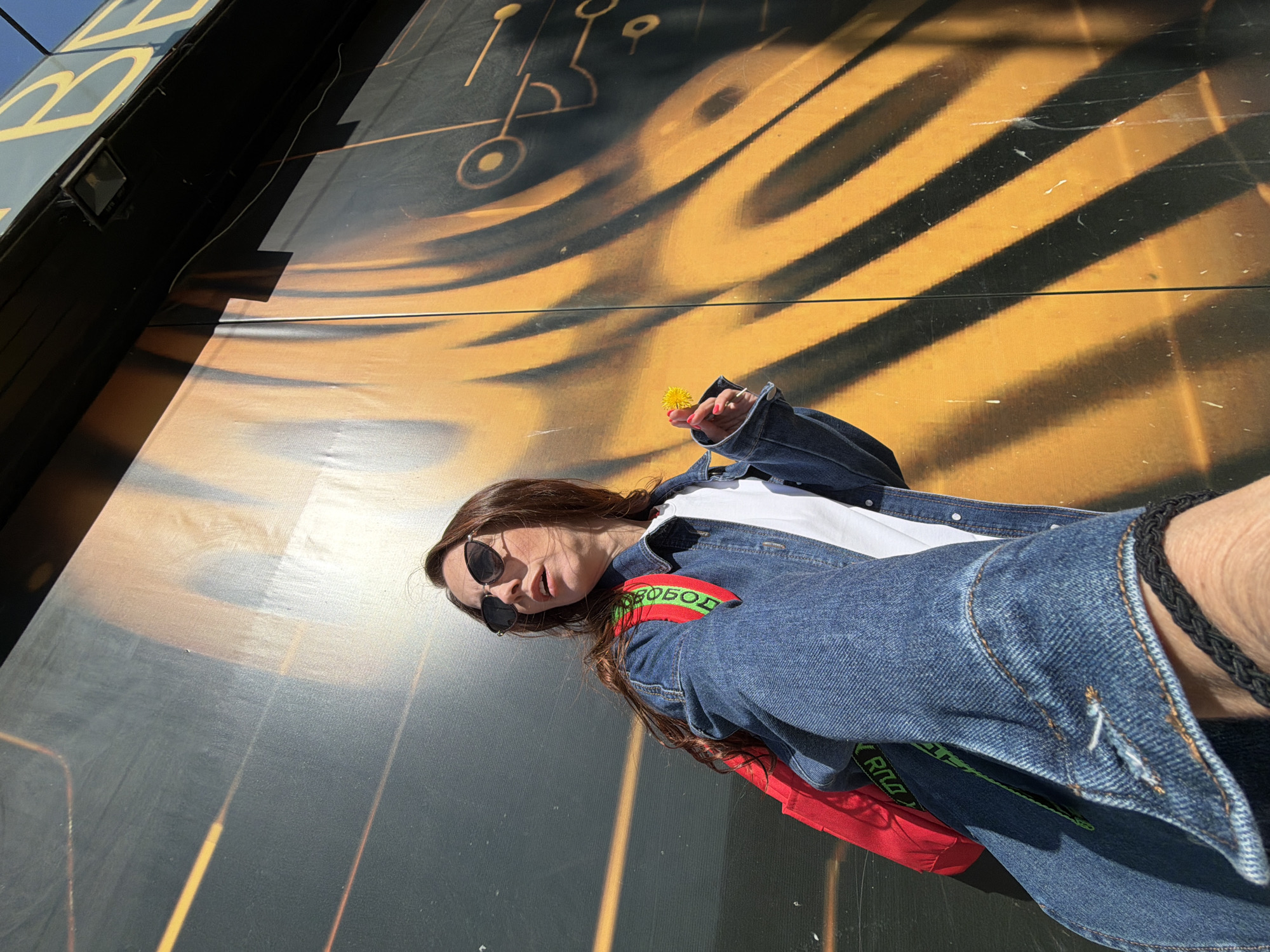 A woman with long, dark hair and sunglasses stands in front of a large mural. The mural features a dark background with an abstract, flowing pattern in orange and yellow, resembling stylized circuitry. The woman is wearing a denim jacket over a white t-shirt and a red backpack with a green strap that has writing on it. She is holding a small yellow flower in her right hand, which is extended towards the mural. A black braided bracelet is visible on her left wrist. The lighting suggests it is daytime, with shadows cast by the mural and potentially the building it's on. The image was taken from a low angle. The context of Birobidzhan, Russia is not visually discernible from the image itself.