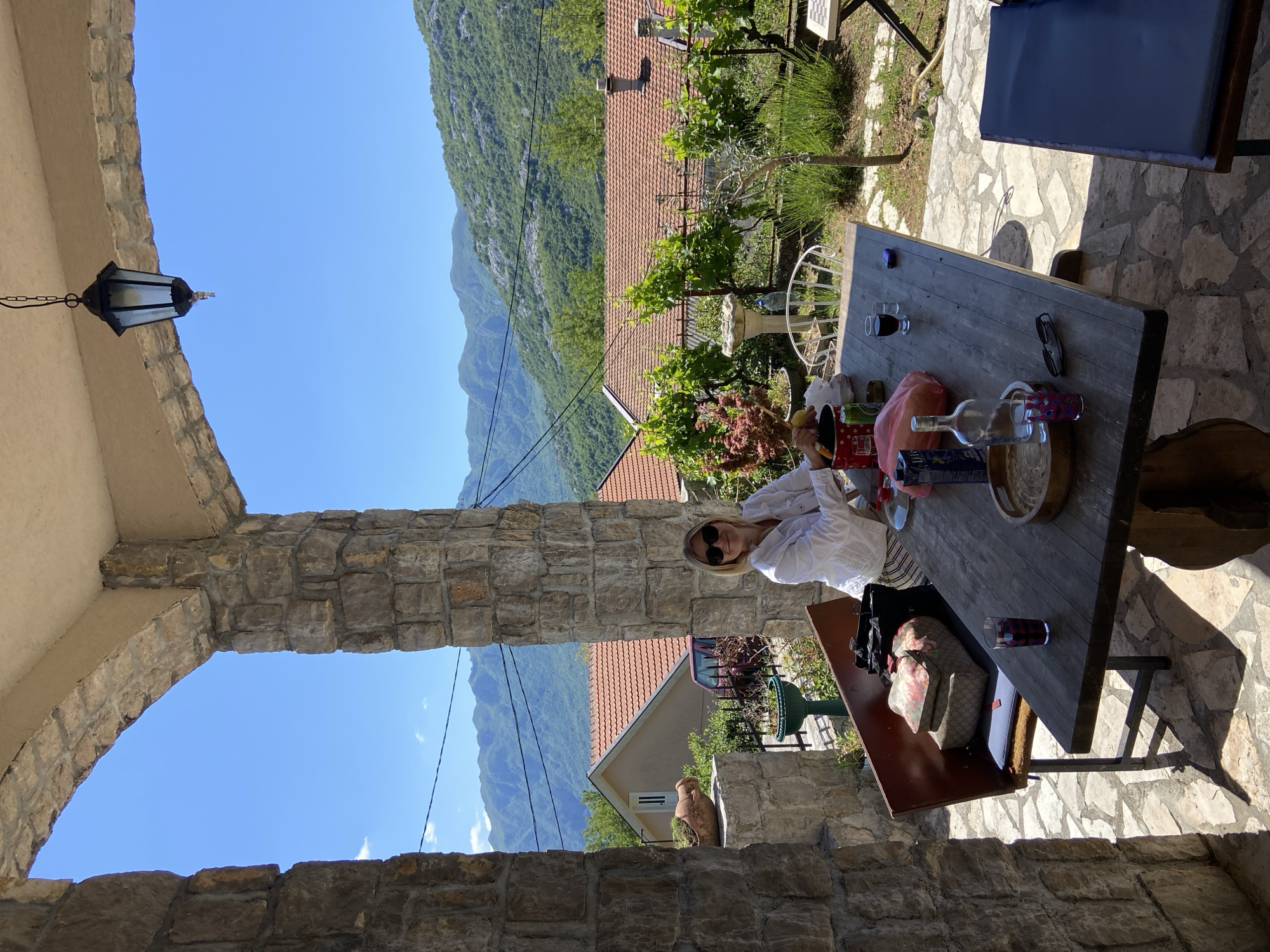 The image, currently rotated 90 degrees clockwise, depicts an outdoor patio scene in Sotonići, Montenegro, overlooking a scenic mountainous landscape. In the foreground, a sturdy stone archway partially frames the view. Hanging from the ceiling of the archway on the left is a traditional black metal lantern.
A woman with blonde hair and sunglasses, dressed in a white shirt, is seated at a rustic wooden table positioned on the stone-paved patio. She is seen from the chest up, leaning slightly forward and looking towards the camera. The table is laden with various items, including wine glasses (one with red liquid), a wine bottle, a clear glass bottle with a white label (text illegible), a checkered glass, a red container or bag, a can (possibly beer or soda), a lemon, and a pair of dark sunglasses. A Gucci handbag rests on a chair next to the table.
Beyond the patio, the landscape unfolds with several houses featuring red tiled roofs, nestled amidst lush green hills and towering mountains under a clear, bright blue sky. Other elements visible on the patio include a chessboard on a stand, a birdbath or fountain, and various potted plants and greenery, contributing to a vibrant garden atmosphere. A large ceramic pot or urn is also visible near one of the houses. The strong shadows cast by objects indicate bright sunlight, suggesting a clear day during the mid-day to afternoon. The overall scene conveys a sense of relaxation in a picturesque, natural setting.