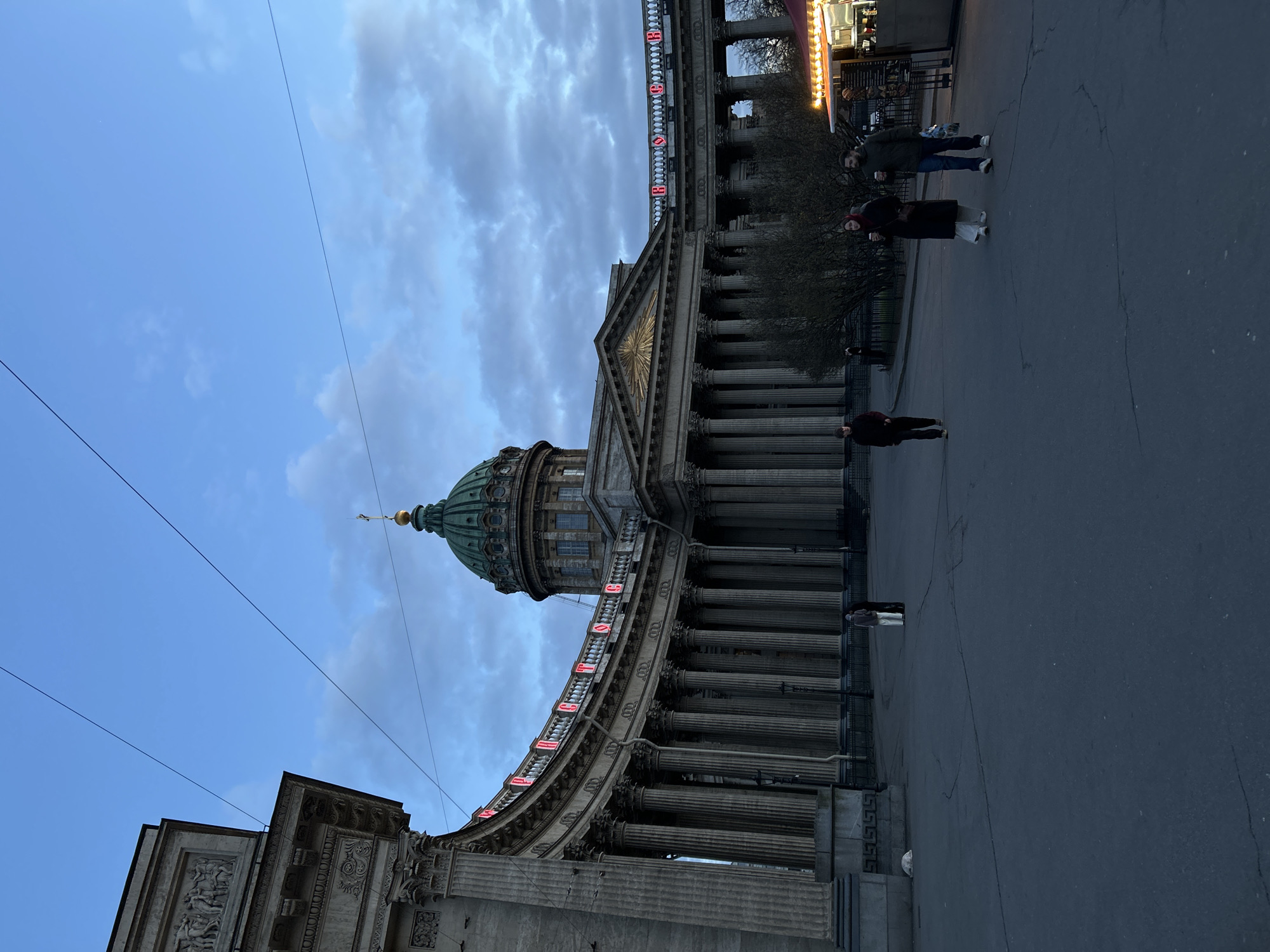 This image captures the grand Kazan Cathedral in Saint Petersburg, Russia, with its distinctive architecture dominating the left side of the frame. The cathedral features a large, dark green dome crowned with a golden finial and is fronted by an imposing semi-circular colonnade of numerous classical columns. A triangular pediment with a sunburst-like relief is visible above one entrance.
Along the balustrade of the colonnade, illuminated red text spells out "С ДНЁМ ВЕЛИКОЙ ПОБЕДЫ" (S DNYOM VELIKOY POBEDY), translating to "Happy Great Victory Day." This text suggests the photo was taken around May 9th, a significant public holiday in Russia.
On the paved street and square in the foreground and midground, several people are walking. Two individuals are seen in the midground, one carrying bags, and another person stands closer to the cathedral's steps. To the far right, a brightly lit food stall with a red awning indicates a public space with pedestrian activity.
The sky above is partially cloudy with patches of blue. Despite the ambient light, the illuminated text and food stall lights suggest it is likely dusk, early evening, or an overcast day. Overhead wires traverse the sky, and cracks are visible on the paved ground. The overall scene depicts a typical urban setting with historical architecture and daily life.