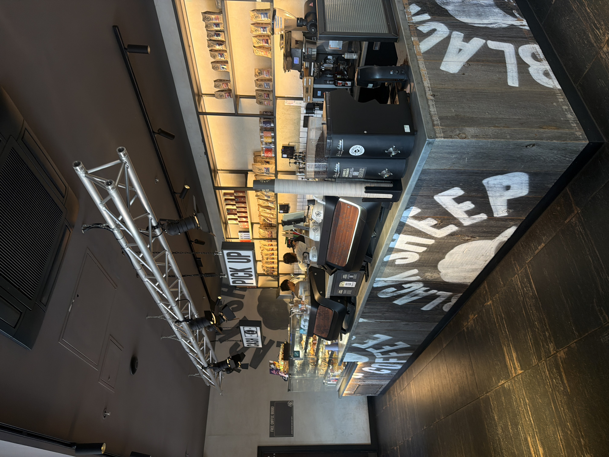 The image shows the interior of a "Black Sheep Coffee" shop, a coffee chain with locations in London. The scene is dominated by a rustic-style counter made of weathered wood panels, adorned with white lettering that reads "COFFEE" and "BLACK SHEEP". Behind the counter, a display case filled with pastries is visible. The back wall features shelves stocked with bags of coffee beans and other products. Overhead, industrial-style lighting trusses are suspended from the ceiling, along with a ventilation system. Two employees are seen working behind the counter, one of whom is interacting with a customer at the display case. The floor is tiled with dark, textured tiles. The overall impression is of a modern and trendy coffee shop.
