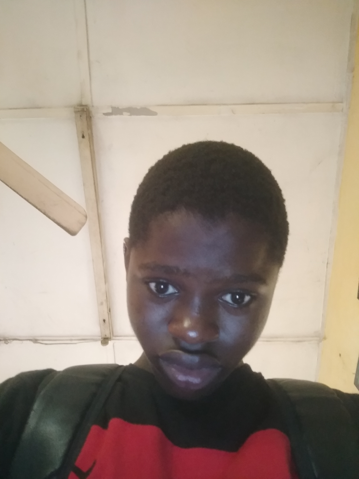 A close-up, low-angle photograph shows a dark-skinned young person in what appears to be an interior space. The individual has closely cropped hair and is wearing a dark shirt with a red design on the front. The background consists of a white ceiling with supporting beams, and part of a ceiling fan is visible in the upper left corner. The image gives no clues to indicate the scene is in Shangisha, Nigeria. No actions or interactions are visible in this static shot.