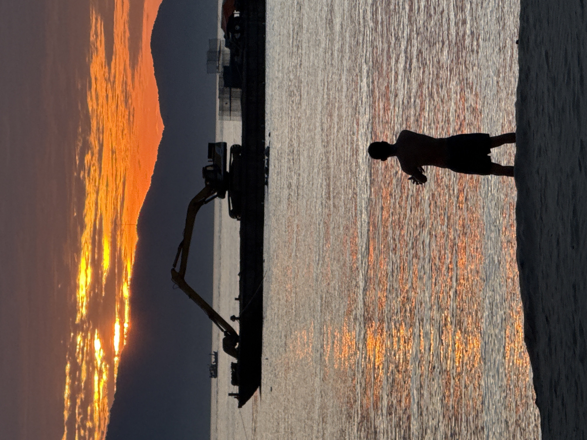 This image depicts a coastal scene in Đà Nẵng, Vietnam, during either sunrise or sunset. The sky, located in the upper half of the frame, features a vibrant display of orange and yellow light radiating from behind dark, irregular cloud formations. Silhouetted mountains with undulating peaks form a dark barrier across the horizon beneath the illuminated sky.
In the middle ground, a large, dark-colored barge with a prominent excavator mounted on its deck is positioned on the water. The excavator's long arm is extended upwards and to the left. Additional unidentified structures are visible on the far side of the barge. To the left of the barge, a smaller, dark-silhouetted boat is visible further in the distance. The surface of the water reflects the orange and yellow light from the sky, creating shimmering streaks and patterns.
In the immediate foreground, a single human figure stands facing away from the viewer, silhouetted against the bright water reflections. The person is partially submerged, standing in shallow water, and appears to be wearing shorts. Their arms are bent at the elbows and held in front of their chest, suggesting a static pose or engagement in a slow, deliberate movement. A narrow strip of dark sand or shoreline is visible beneath the person in the bottom foreground.