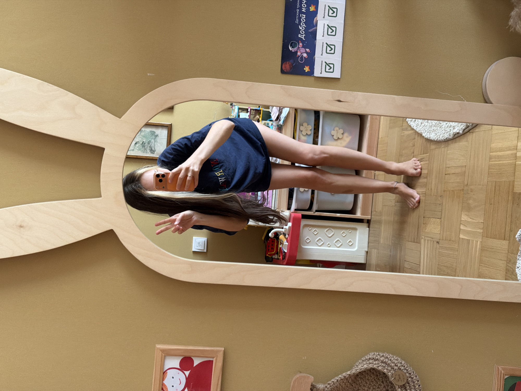 A young woman with long, dark hair, some of which is blonde, takes a mirror selfie in a room. She is wearing a dark blue t-shirt and no pants. She is standing barefoot on a wooden floor. In her right hand, she holds an orange smartphone, which is pointed at the mirror. Her left hand is raised, and she is making a peace sign.
The mirror is framed with light-colored wood and has a stylized bunny ear shape at the top. Reflected in the mirror, a toy kitchen set with red and white elements is visible, along with storage bins with flower-shaped embellishments. On the wall to the left of the mirror, a framed drawing of a red figure with a smiling face is hanging. To the right of the mirror, a chart with a drawing of an astronaut and Russian text that reads "Доброй ночи" (Good night) is attached to the wall. The wall is painted a muted gold color, and the floor is made of wood parquet. The room appears to be a child's bedroom or playroom. The image was taken in a residential apartment located in Khimki, Russia.