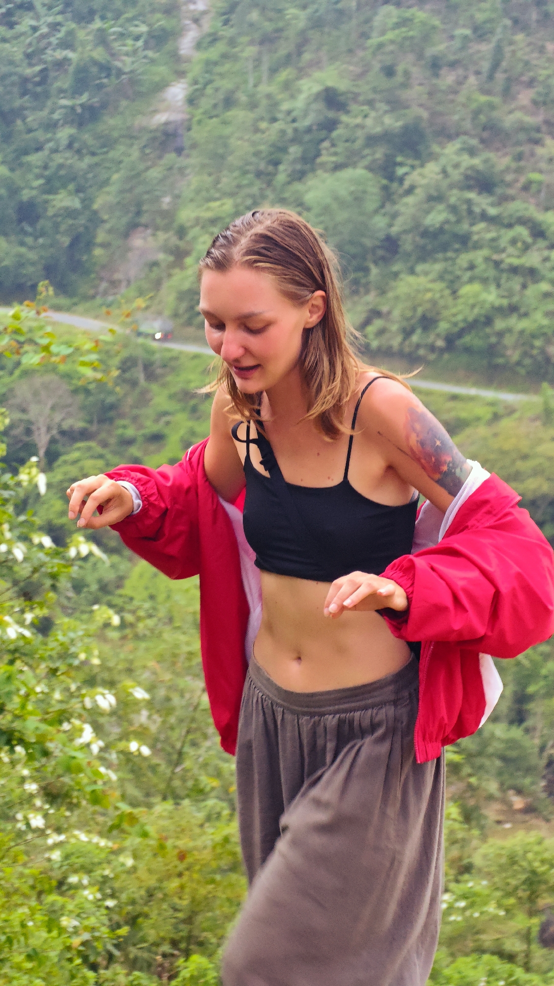 A young woman is captured in a vertical, outdoor shot, set against a backdrop of lush green hills and dense vegetation. She has light brown hair, fair skin, and her eyes are mostly closed with a soft smile, suggesting a moment of joy or engagement. She is wearing a black spaghetti-strap crop top, revealing her midriff, and loose-fitting, dark grey pants. A vibrant red jacket is draped over her shoulders and upper arms, with her hands partially out of the sleeves. A colorful tattoo is visible on her left upper arm. Her hands are raised slightly, fingers gently curled, as if in motion or interacting with something out of frame.
The background features multiple layers of green foliage, indicating a natural, hilly, or mountainous landscape. A narrow road with a small, indistinct white vehicle can be seen in the mid-ground on the left, winding through the greenery. The lighting suggests it is daytime, likely morning or afternoon, with natural light illuminating the scene. The overall atmosphere appears tranquil and natural, consistent with the location context of Nậm Dịch Commune, Vietnam. There is no visible text in the image.