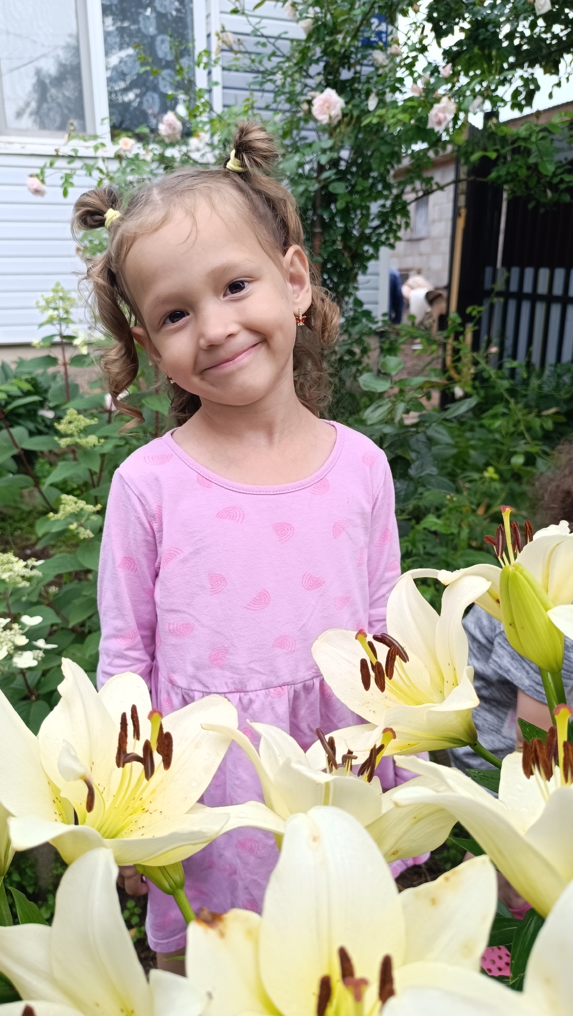 A young Caucasian girl with curly brown hair in pigtails, wearing a pink long-sleeved dress, smiles at the camera. She is standing in a garden in front of a light blue house. Several large white lilies are in the foreground, obscuring part of the girl's lower body and the immediate surroundings. The background shows a white house with a window, green foliage, and a dark fence. The overall lighting suggests it is daytime. No specific details about Karsha, Russia are visible or implied in the image.