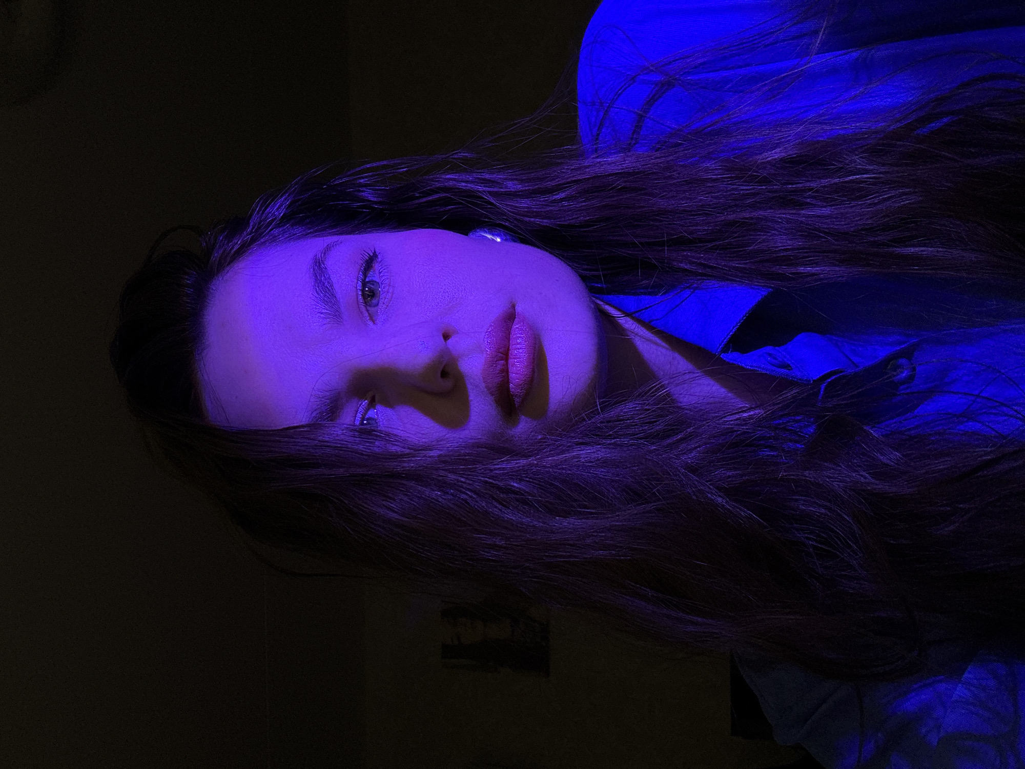 The image shows a young woman's face lit with a strong blue/purple light. Her long, dark hair is partially obscuring her face. She wears what appears to be a blue shirt. The background is dark. There is no evidence in the image that places it in Chaikovsky, Russia or any other location.