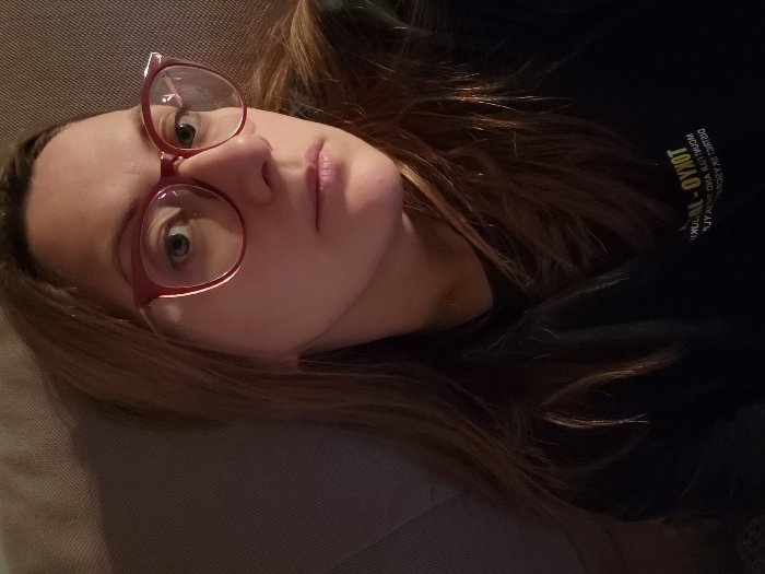 A woman with light skin and brown hair is seen lying down. She wears red-framed glasses. Her eyes are visible. She is wearing a black shirt. Some lettering is visible on the upper right of the shirt. The backdrop is a light beige. The composition is a close-up shot.
