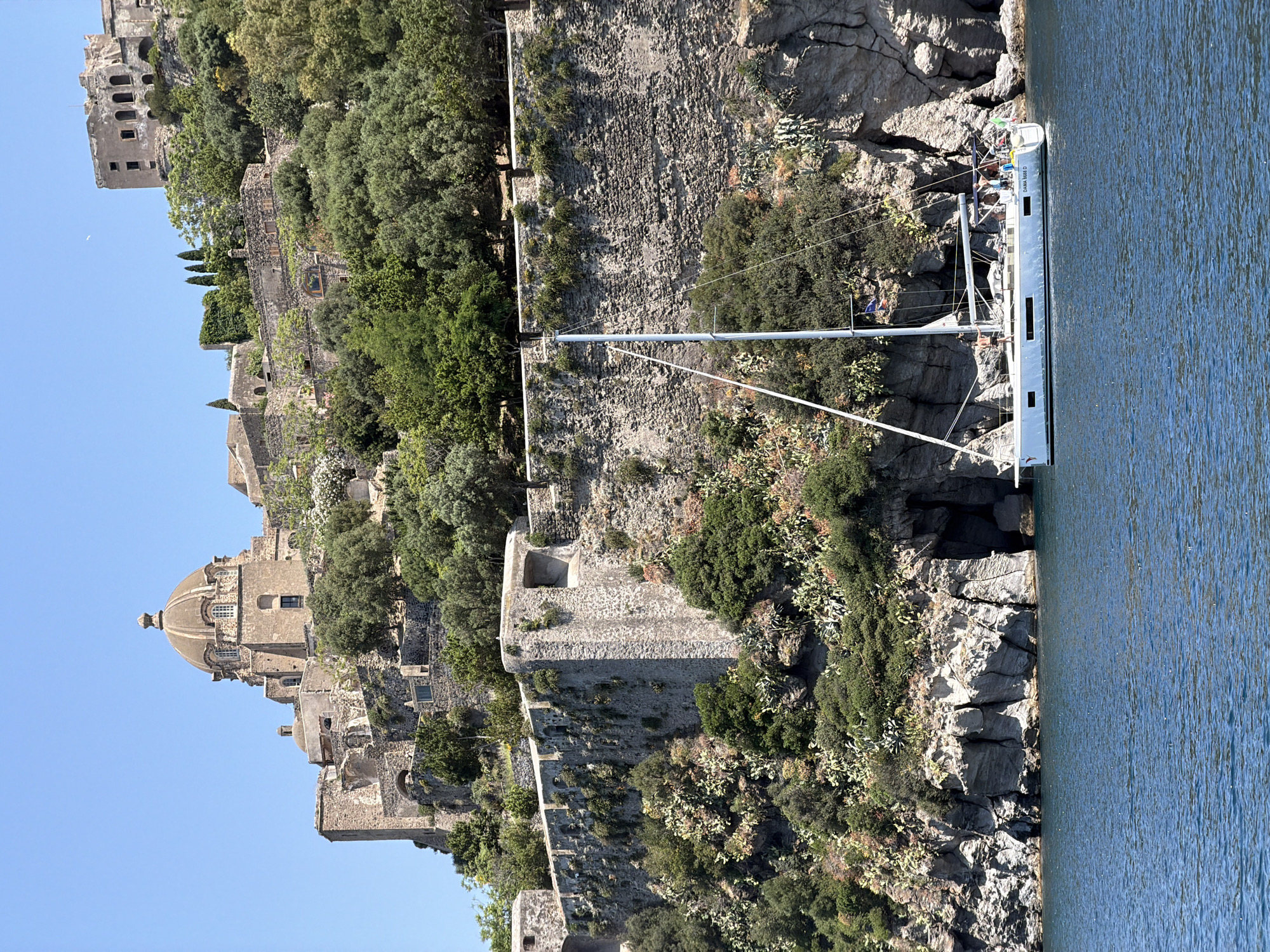 A vertical, eye-level shot shows the Castello Aragonese in Ischia, Italy, along with a sailboat docked nearby.
The left side of the frame is dominated by the Castello Aragonese, a medieval castle complex rising up the side of a rocky, terraced hillside. The architecture is characterized by stone walls, ramparts, and buildings of varying heights. A prominent dome-shaped roof is visible near the lower portion of the castle, with other structures and walls stretching upwards towards the top left corner of the frame. Green vegetation, including trees and bushes, grows among the stone structures.
On the right side of the frame, a white sailboat is docked against a rocky cliff. The boat's mast extends upwards, and people are visible on board. The cliff face is textured with gray rocks and interspersed with green plants.
The water of the Mediterranean Sea fills the lower right portion of the frame, reflecting the sky. The sky is clear and blue in the background behind the castle.