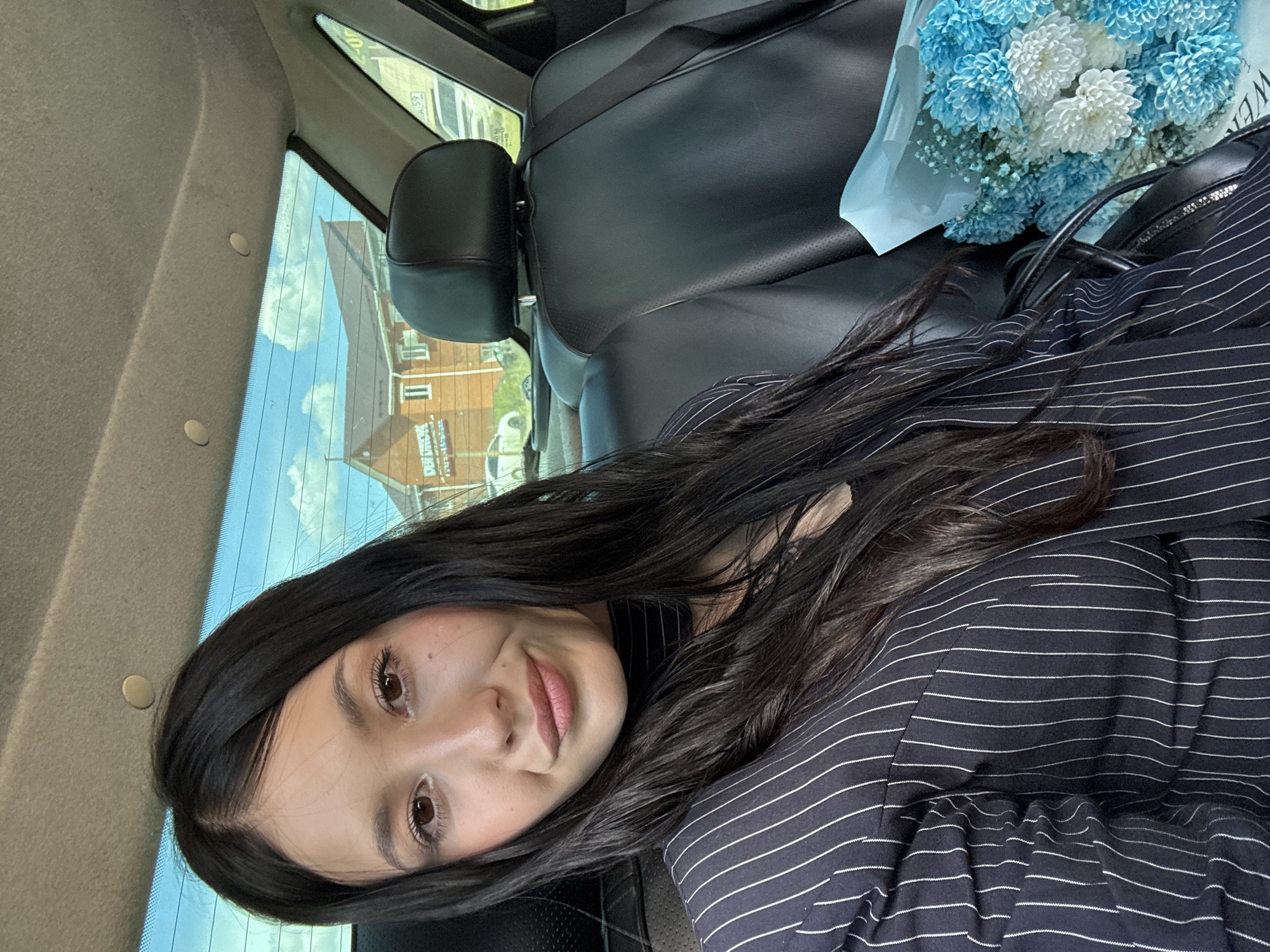 A young woman with long, dark hair is taking a selfie in the back seat of a car. She is wearing a dark pinstripe blazer and a black scarf. A bouquet of blue and white flowers is on her right. The car's interior is visible, with black leather seats and a headrest. Through the rear window, a two-story building with a dark roof and white siding is visible, along with trees and a blue sky with clouds. This building is identified as a residential building located on Privokzalnaya Street in the city of Chaikovsky, Russia.
