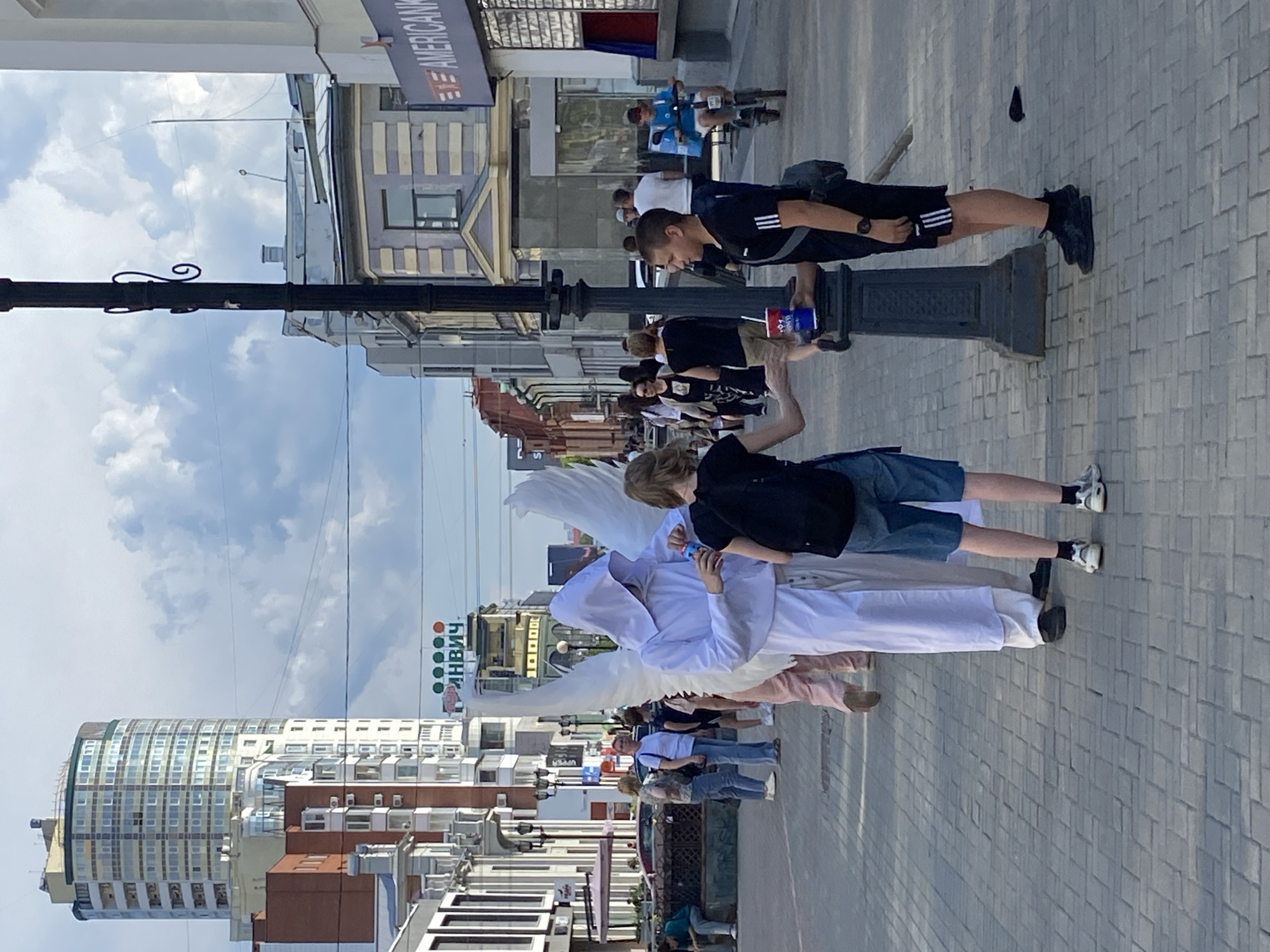 In Yekaterinburg, Russia, on a paved pedestrian street, a person dressed as an angel with large white wings and a hooded white robe is interacting with two young people. The angel, whose face is obscured by the hood, is holding a blue cup with a red lid and appears to be offering it to the person on the right, a young male wearing a black t-shirt with white stripes and black shorts. This male is reaching for the cup, holding a similar red and blue cup in his other hand. To his left, a young female wearing a black t-shirt and blue shorts is partially visible, her left hand outstretched towards the angel. In the background, other pedestrians are walking along the street, and buildings line both sides. One prominent building features a curved facade and multiple windows. A street lamp with decorative elements stands between the angel and the young male. The scene is illuminated by daylight under a cloudy sky.