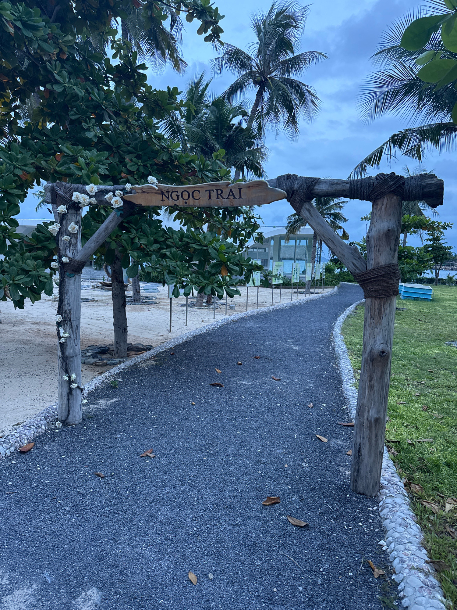 The image shows a rustic wooden archway with a sign that reads "NGỌC TRÀI". The archway is decorated with white artificial flowers and rope. A dark gray gravel pathway curves to the right, bordered by small white pebbles on the left and green grass on the right. Palm trees and other lush green foliage surround the pathway. In the background, a modern building with glass windows and several informational displays is visible. A light blue object, possibly a lounge chair, sits on the grass to the right. The scene appears to be a resort or a similar tourist destination, likely in Nha Trang, Vietnam, given the tropical vegetation and architectural style. The sky is overcast, suggesting it is either early morning or late evening. There are no people visible in the image.