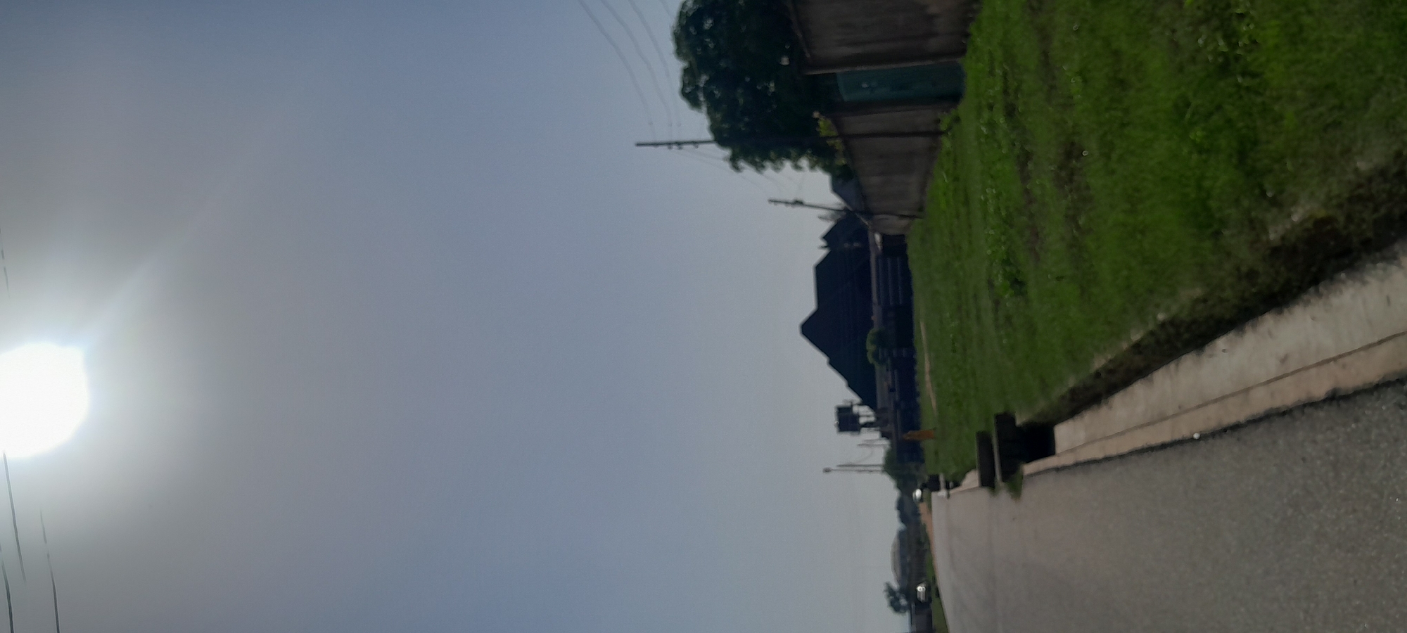 The image depicts a street scene in Jalingo, Nigeria, on a bright, sunny day with a clear sky. The perspective is from the side of an asphalt road, looking down its length. To the left, the road curves slightly, with a few vehicles visible in the distance. A concrete drainage channel runs alongside the road on the right, bordered by a strip of green grass.
Further down the road, on the right side, a large, dark-roofed building with a complex, angular design stands prominent. Adjacent to this building is a tall concrete wall, likely a boundary fence, with a green gate visible. Behind the wall, lush green trees are abundant, suggesting a well-vegetated area. A utility pole with power lines is also visible. In the far distance, more buildings and trees can be observed. The sun is high in the sky, casting bright light and creating some overexposure in the upper part of the image. The overall impression is one of a residential or commercial area under development or with established infrastructure.