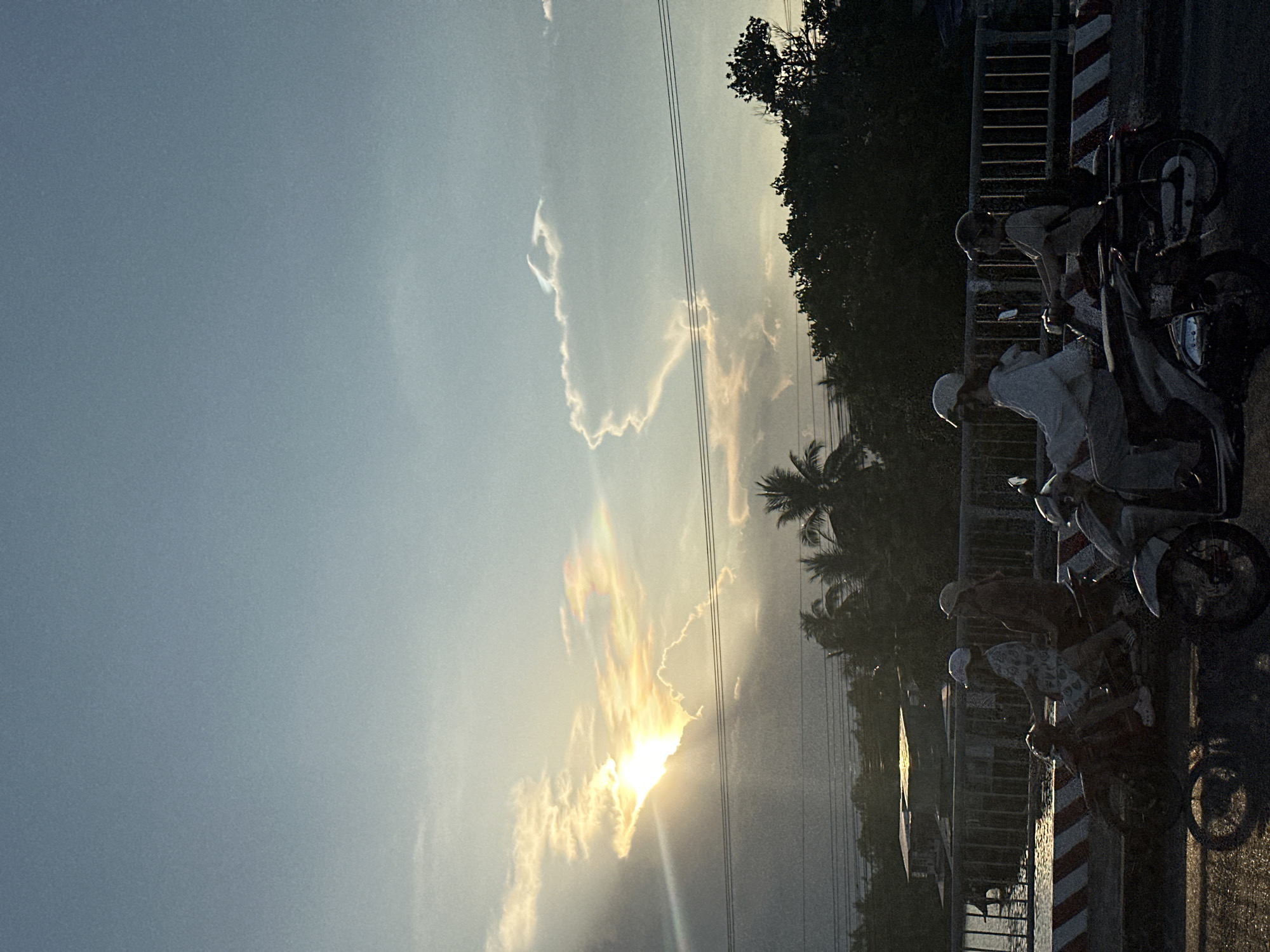 A vertical photograph shows a sunset over Nha Trang Ward, Vietnam, with motorcycles on the right side of the image. The sun is at the bottom center of the image, obscured by clouds. The sky is blue, with a few clouds scattered throughout. Several electrical wires run horizontally across the image. Palm trees and other vegetation are visible on the right side. Four motorcycles are parked on the right side of the image, along with a bridge railing. People wearing helmets sit atop some of the motorcycles. The scene appears to be taken during dusk, as the light is dim.