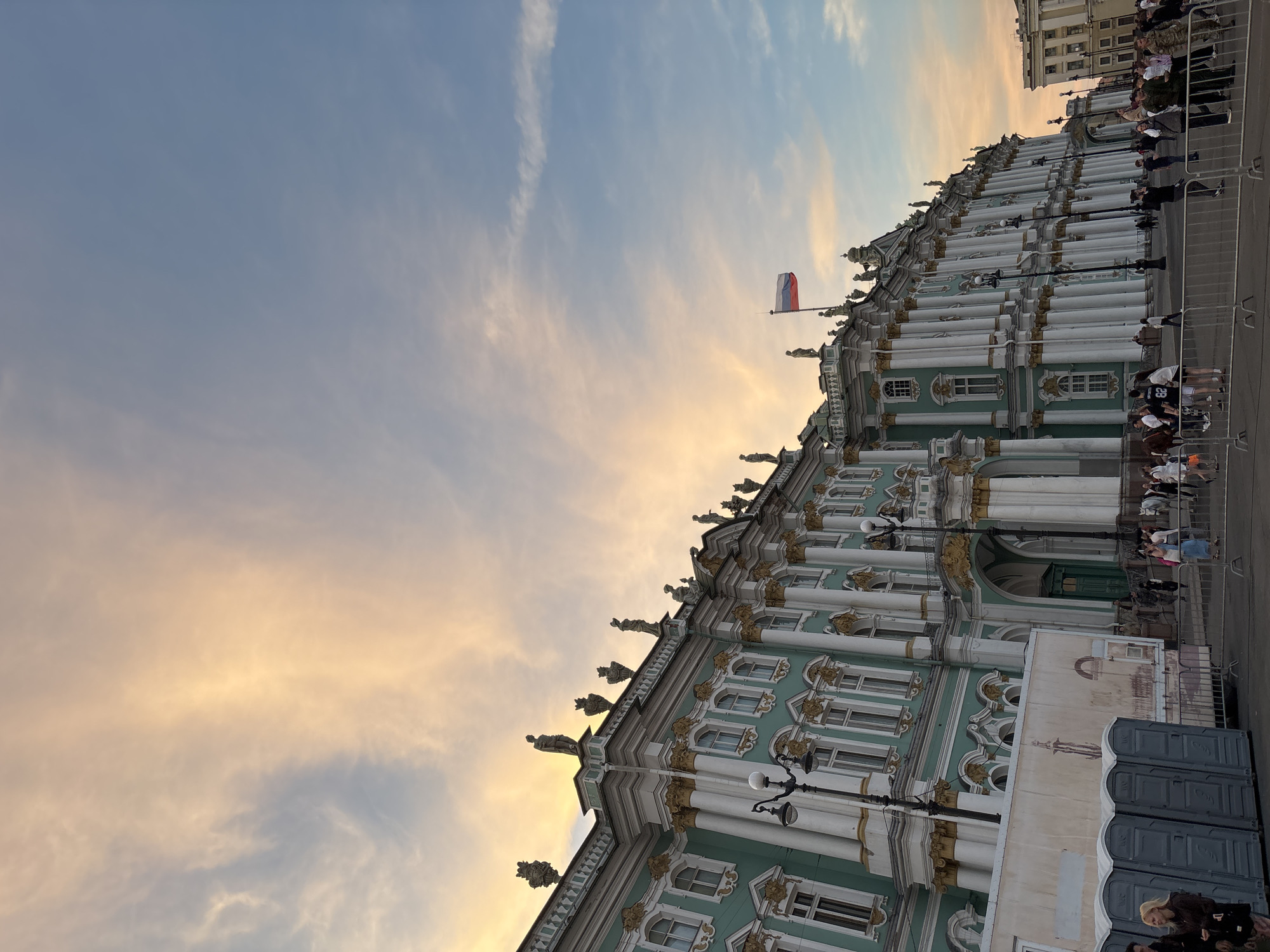 The image depicts the Palace Square in Saint Petersburg, Russia, with the Winter Palace as the dominant structure. The sky above is a canvas of soft, warm colors, transitioning from a peachy orange glow to a pale blue, suggesting either sunrise or sunset. Wispy clouds are scattered across the sky, adding texture and depth.
The Winter Palace, with its distinctive mint green facade and ornate gold detailing, stands majestically. Its classical architecture is emphasized by rows of tall, white columns and numerous windows. Statues adorn the rooftop, silhouetted against the colorful sky. A Russian flag flies from a flagpole atop the building, a clear indication of the location.
In the foreground, a crowd of people are gathered in the square, some walking, some standing and observing. Their casual attire suggests it's a public space accessible to tourists and locals. Several portable toilets are visible on the left side of the image, along with temporary fencing, possibly set up for an event or to manage visitor flow. Other buildings in the background, though less detailed, contribute to the urban landscape. The overall atmosphere is one of a vibrant public space bathed in the gentle light of the sky.