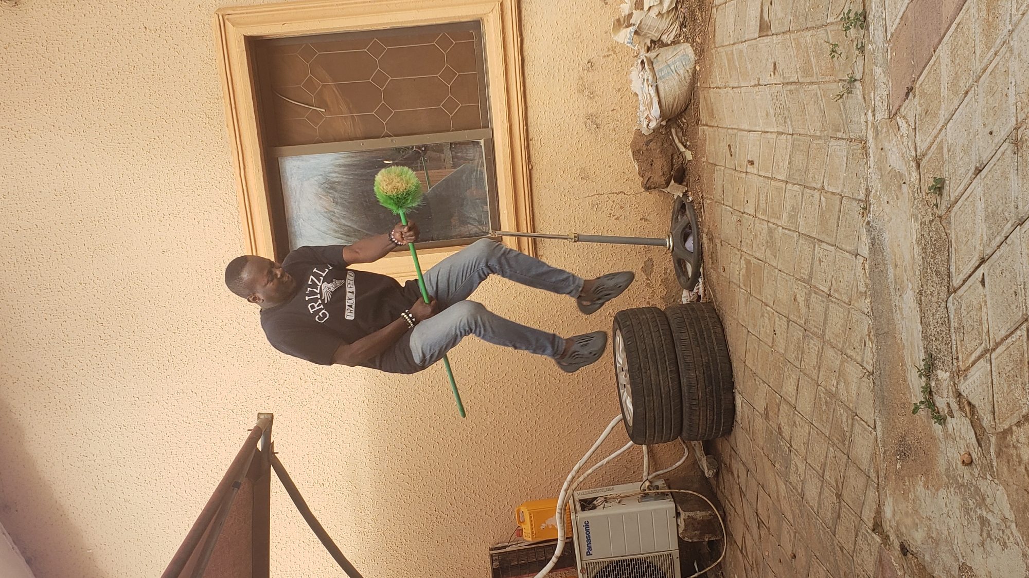 This outdoor photograph, set in Sabon Tasha, Nigeria, shows a man posing next to a textured light brown building wall under bright daylight. The man, with short dark hair, is dressed in a black t-shirt emblazoned with "GRIZZLY TRACK & FIELD," grey jeans, and dark, textured slip-on shoes. He holds a vibrant green and yellow duster, which appears to be propped on a metal bar extending from a black weight plate on the ground. His body is captured in a dynamic, mid-air pose with his feet off the ground, suggesting either a jump or a playful suspension, likely using the duster and weighted bar for support.
Behind the man, the building features a window with a wooden frame and a decorative, patterned grille. The ground is a mix of diamond-patterned concrete paving and areas of dirt with sparse greenery. To the man's right, two car tires are stacked horizontally next to two outdoor air conditioner units; one unit clearly displays the "Panasonic" brand. A yellow plastic container and several light-colored sacks are also visible on the ground near the wall. The strong sunlight indicates a clear day, casting distinct shadows and illuminating the scene of this everyday outdoor environment.