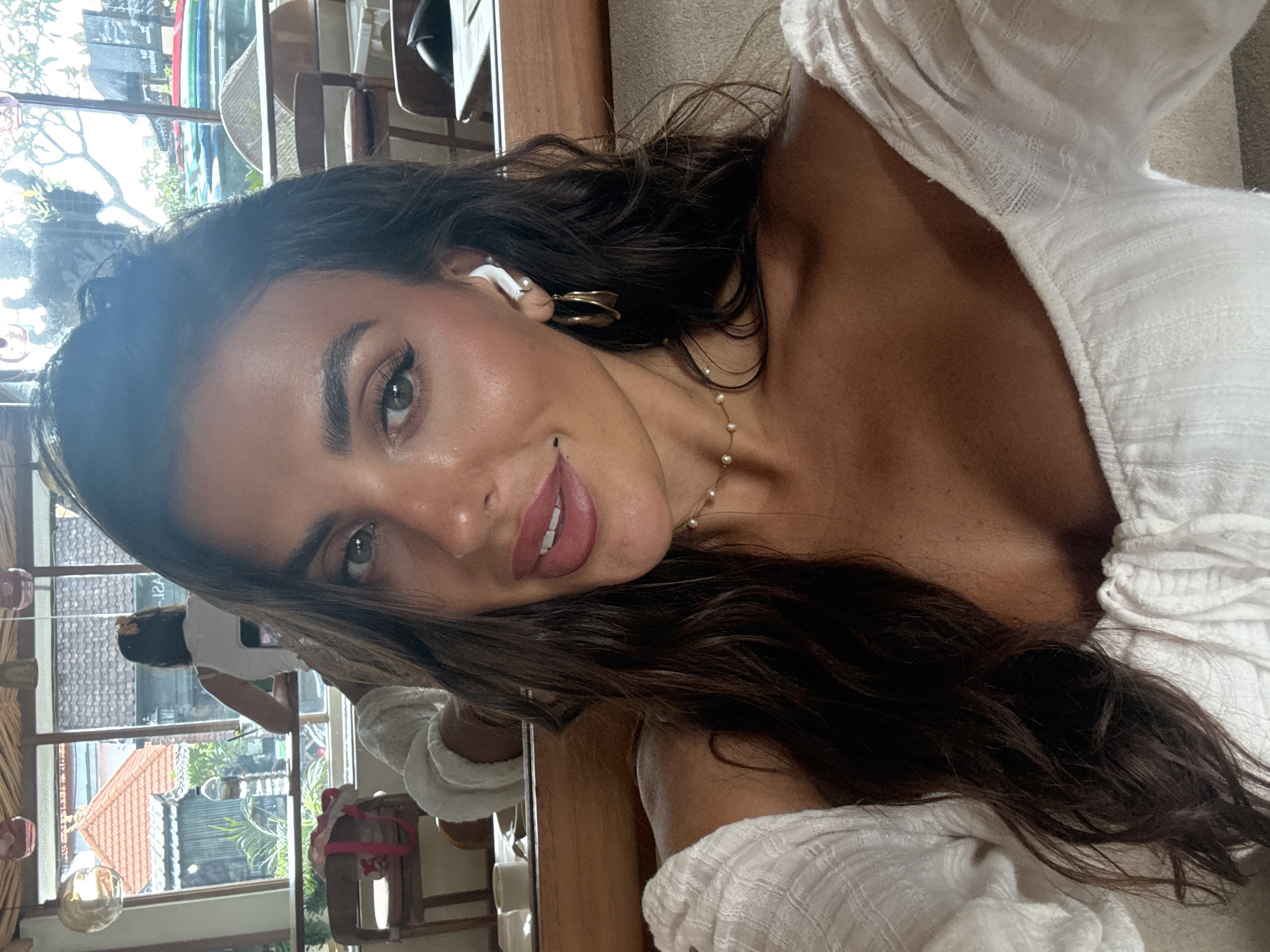 A woman poses for a selfie indoors, possibly in a cafe or restaurant in North Kuta, Indonesia. She has long, dark, wavy hair and is wearing a white, off-the-shoulder top. She is also wearing gold hoop earrings, a pearl necklace, and an earpiece in her left ear. Behind her, a window reveals glimpses of the outdoors, including trees, a building with a red roof, and various objects, including what looks like furniture and other individuals.