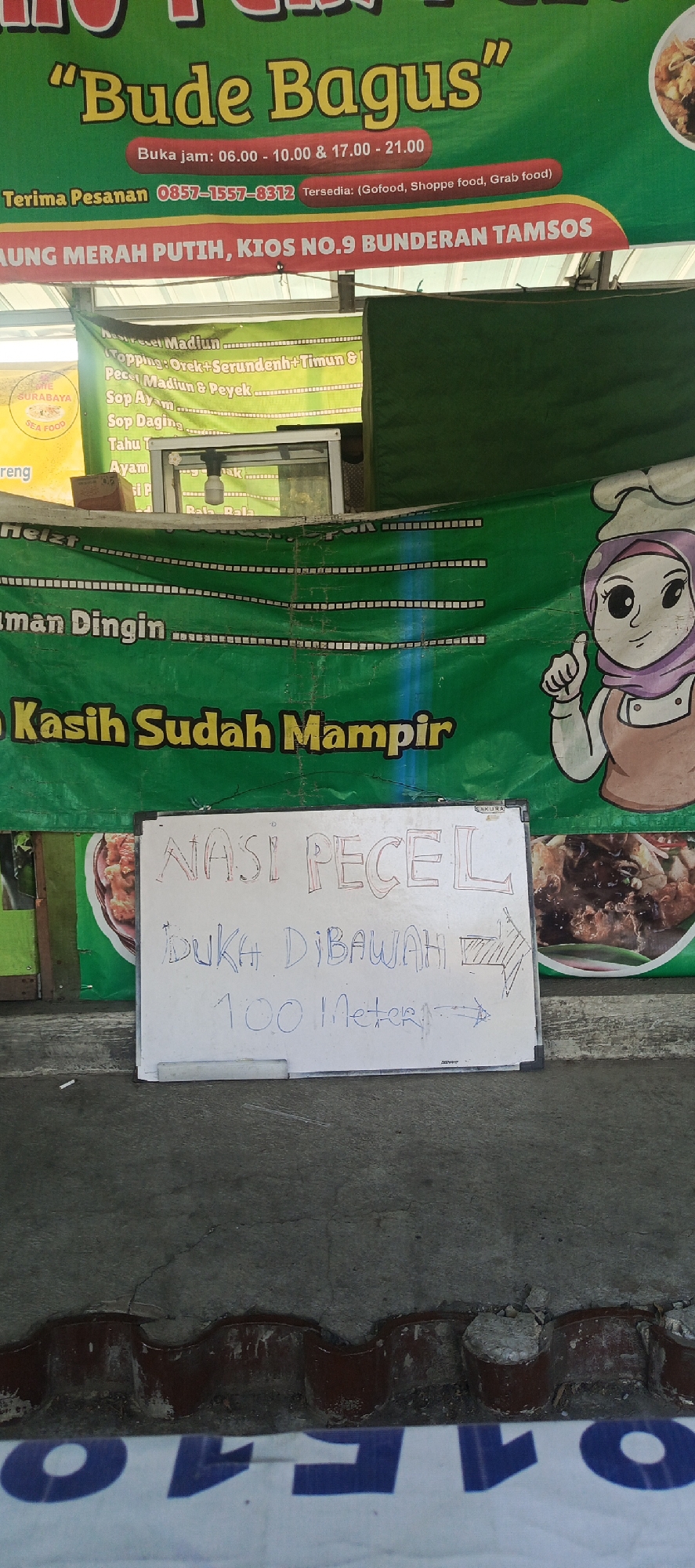 The image shows a storefront for a food business named "Bude Bagus" in Sukaresmi, Indonesia. The business appears to be a street food vendor or small eatery. The main signage is green with yellow lettering and features the business name, operating hours (06.00 - 10.00 & 17.00 - 21.00), and a phone number (0857-1557-8312) indicating availability for delivery services like GoFood, Shopee Food, and Grab Food. There is also a banner advertising menu items such as "Nasi Merah Putih," "Kios No. 9 Bunderan Tamsos," and various toppings and sides including "Pecel Madiun & Peyek," "Sop Ayam," "Sop Daging," "Tahu T," and "Ayam."
In the foreground, a whiteboard displays a handwritten message: "NASI PECEL BUKA DIBAWAH 100 Meter →". This indicates that the Nasi Pecel specialty dish is available at a location 100 meters away, with a directional arrow.
The environment appears to be outdoors, with the storefront set up along a street or walkway. The background includes a green banner with a cartoon illustration of a chef, and elements of the surrounding structures suggest a commercial or semi-urban setting. The ground in the foreground is a textured concrete surface.