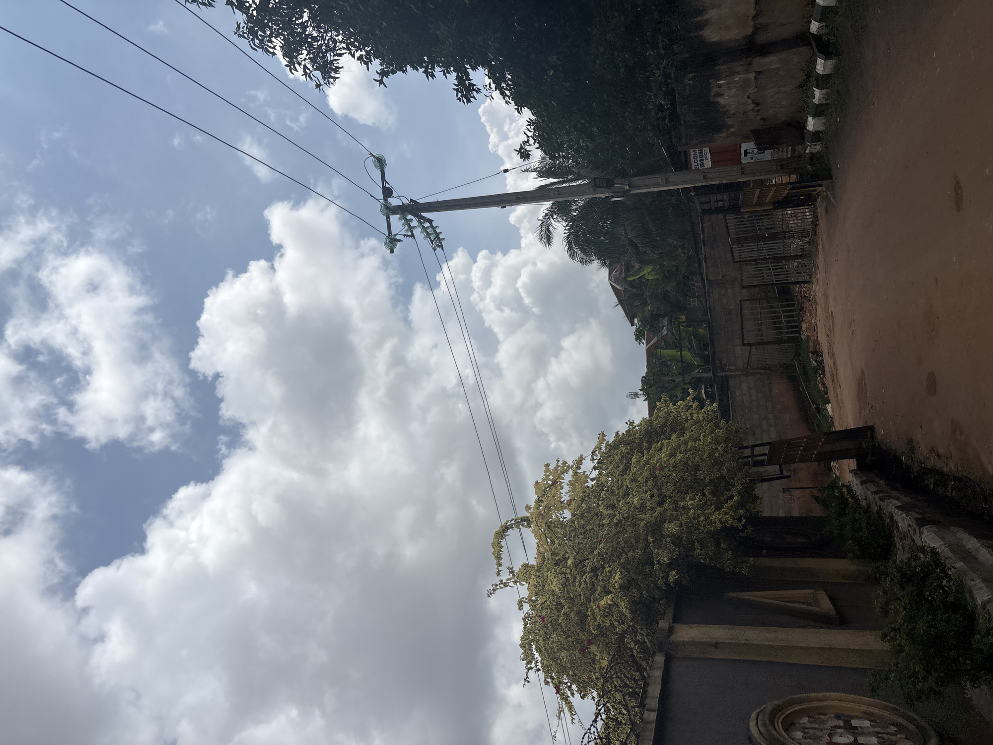 This is a vertical, low-angle shot of a residential street in Nkpor, Nigeria, under a cloudy sky. The scene is dominated by a utility pole with multiple power lines stretching across the frame. Lush green trees frame the top and right sides of the image, with some flowering vines cascading down a wall on the right. A dirt road runs along the right side, bordered by a low wall and a fence. Buildings with visible fences and metal gates are present, suggesting a residential or commercial area. A sign with text is partially visible on a building near the utility pole. The clouds are dense and white, indicating potentially overcast or rainy weather, common in tropical regions. The time of day appears to be daytime, likely afternoon, given the bright, diffused light. The overall impression is one of a typical street scene in a tropical urban or semi-urban environment.