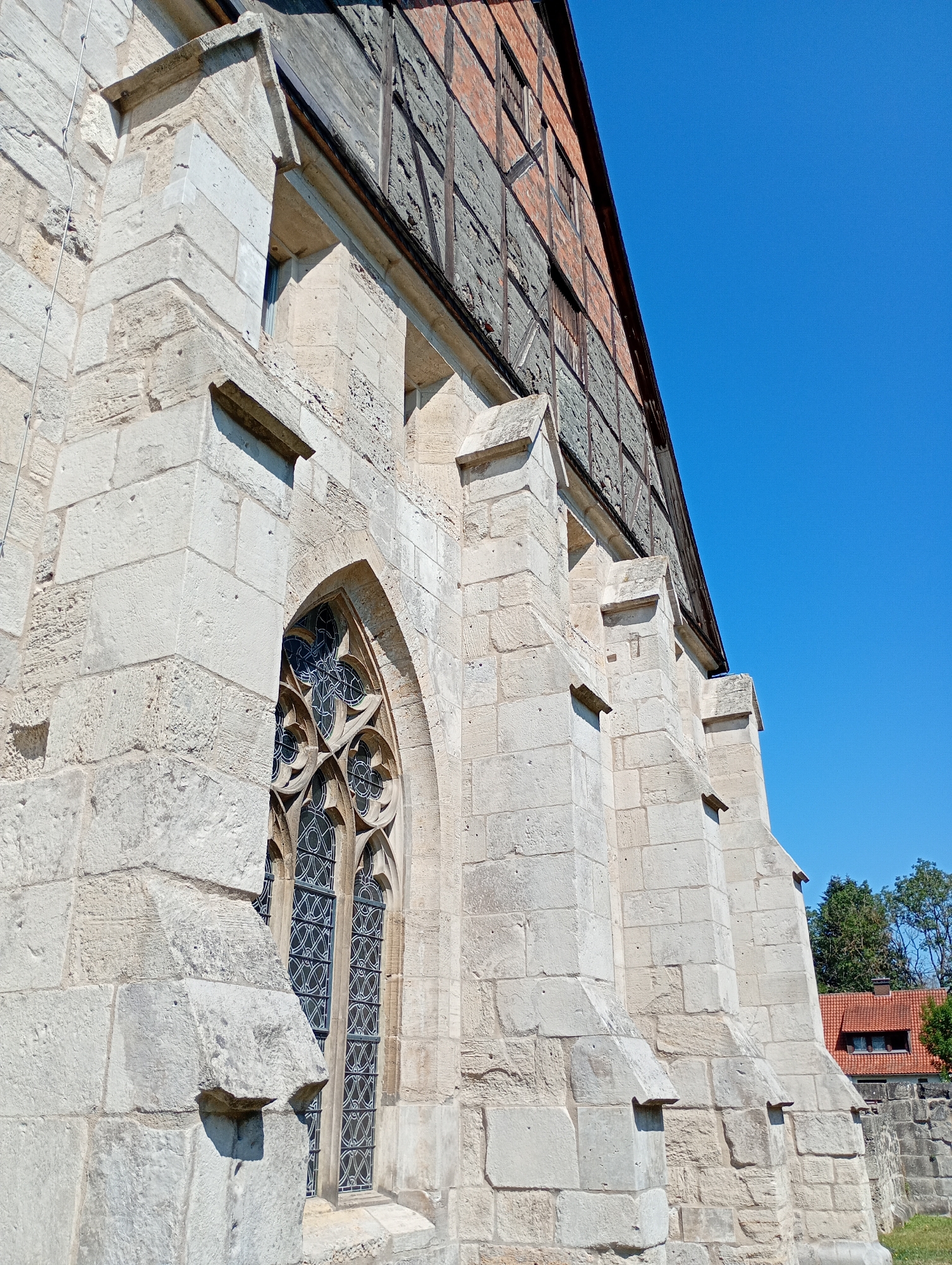 The image shows the exterior of the Johanniskirche (St. John's Church) in Walkenried, Germany. The building is constructed with large, rough-hewn light-colored stone blocks forming the lower portion, with visible buttresses and decorative stonework around a Gothic arched window. The upper part of the building displays a timber-framed facade with dark wood beams and reddish-brown brick infill. The clear blue sky occupies the upper right portion of the frame. In the background, a red-tiled roof of another building and some trees are visible. The scene appears to be a sunny day with harsh shadows cast by the architectural elements. There are no people or animals visible in the image.