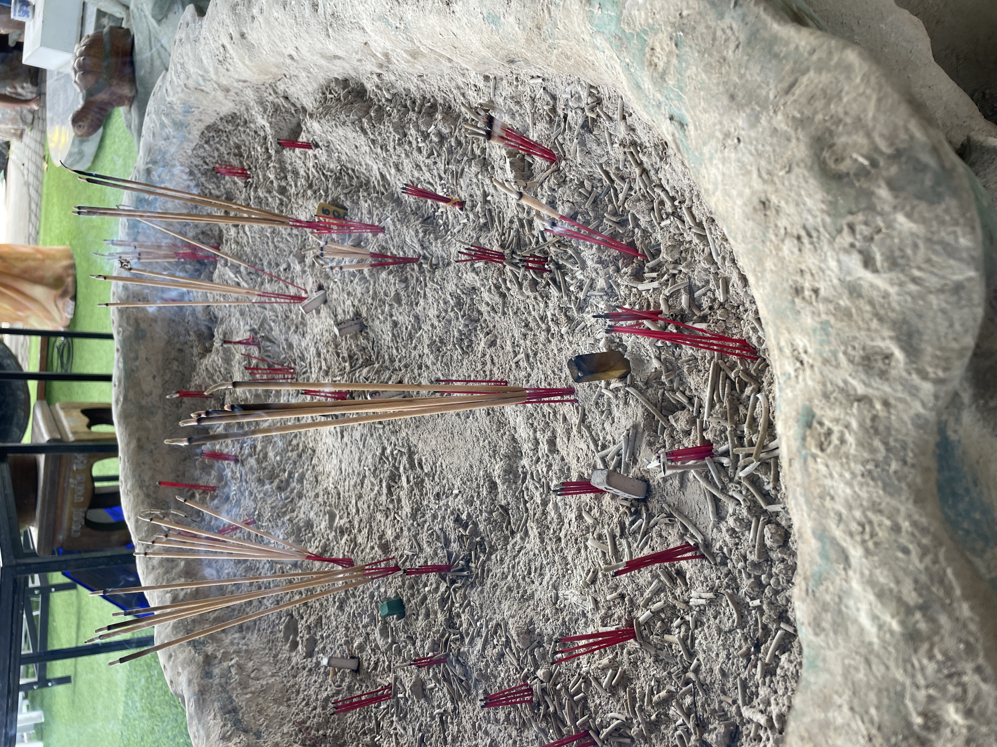 In Tambon Na Chom Thian, Thailand, a large, weathered stone incense burner, filled with sand and the remnants of burnt incense sticks, is depicted. Numerous unburnt incense sticks, many with red bases, stand upright in the sand, while others lie scattered amongst the ashes. The sand is textured with both the fine particulate matter of ash and the coarser grains of sand, interspersed with the cylindrical remains of burnt incense. A subtle wisps of smoke rise from some of the burning incense sticks, indicating their recent use. The surrounding environment is partially visible, showing a green grassy area, a table with a metal frame, and what appears to be a small decorative turtle statue. The lighting suggests an outdoor setting, likely during the daytime.