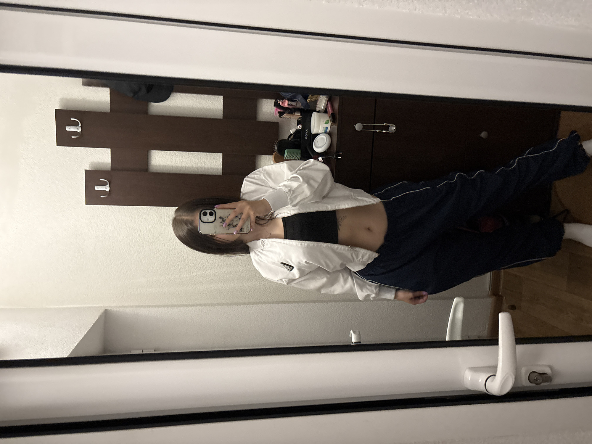 An individual takes a mirror selfie indoors, obscuring most of her face with a smartphone held in her right hand. She has long, dark hair and light purple nail polish. She is wearing a black crop top, a white unzipped bomber jacket, dark blue track pants with a white stripe, and white socks. A navel piercing and a small tattoo on her left ribcage are visible. The reflection shows a domestic setting with white walls and a wooden floor. To the left of the individual is a white door frame with a white lever handle and a key inserted into the lock. To her right is a dark wooden coat rack with two silver hooks (one double, one single), on which a dark cap hangs. Below the coat rack is a dark wooden dresser holding various personal items, including hairbrushes, a white jar, a green-labeled jar, and a white electronic device. There is no visual information within the image to identify the location as Polena, Bulgaria.