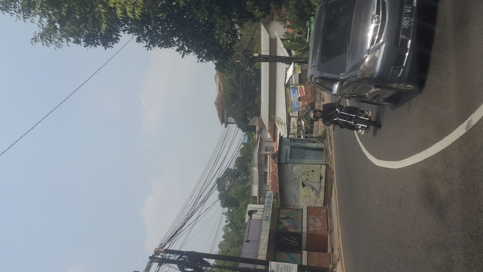 The image captures a street scene in Cilaku, Indonesia. A gray car is parked on the right side of the road, while a person on a motorcycle is riding in the lane. The motorcycle rider is wearing a dark jacket and a helmet. The road is paved and has a white line marking the lane. On the left side of the road, there are several buildings, including a structure with a sign that reads "BAKSO CIHARAS". There are also power lines overhead, characteristic of an urban environment. The sky is bright and clear, indicating daylight. The background shows trees and more buildings, suggesting a populated area within the town.