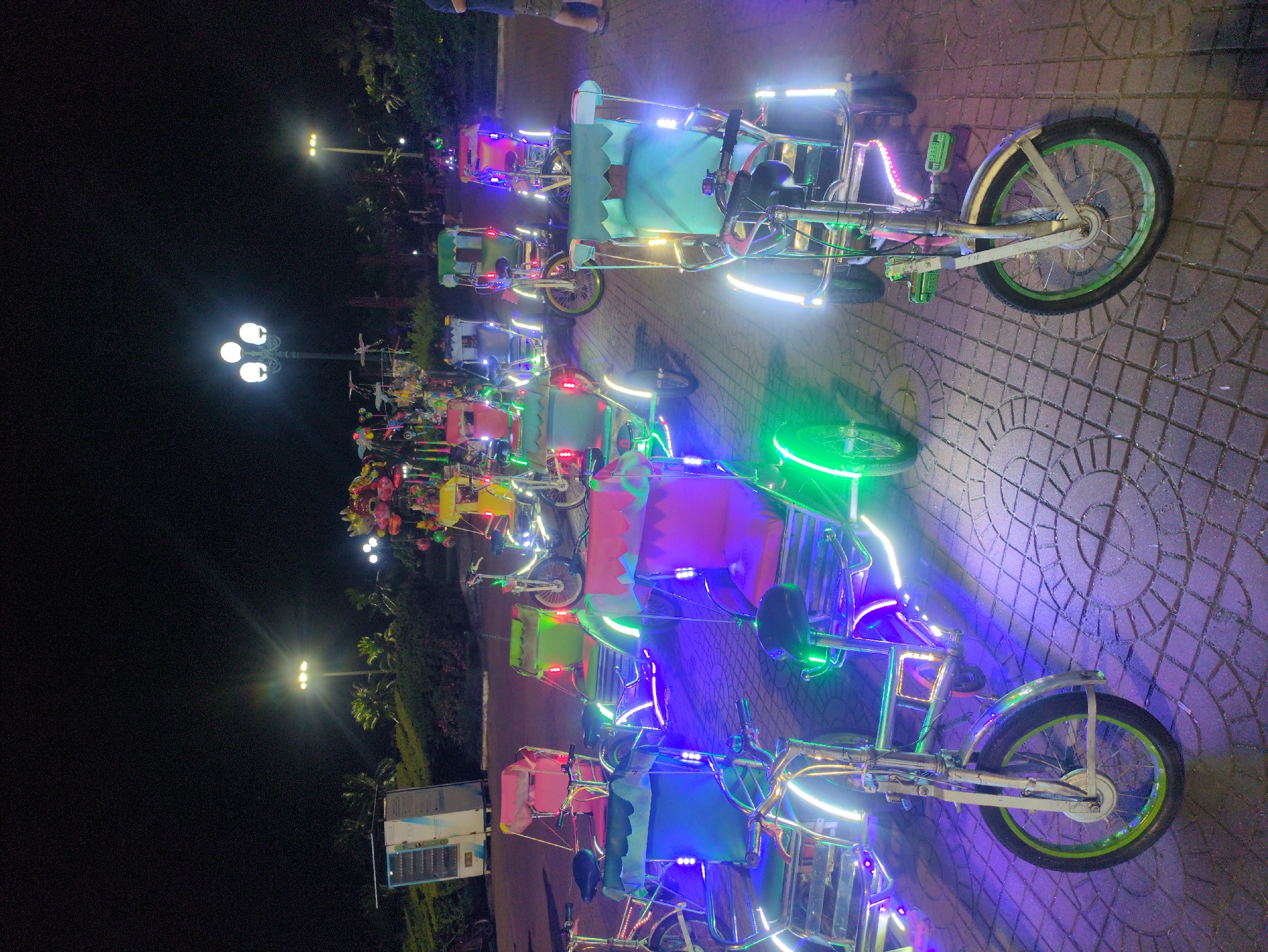 The image shows a row of decorated quadricycles on a paved surface, likely in the city of South Quy Nhon Ward, Vietnam. The vehicles are illuminated with colorful lights, including green, blue, pink, and white. Each quadricycle features a canopy-like structure above the seating area. In the background, there are streetlights and trees. The overall setting suggests a public park or recreational area at night. The vehicles appear to be lined up for rent or use.