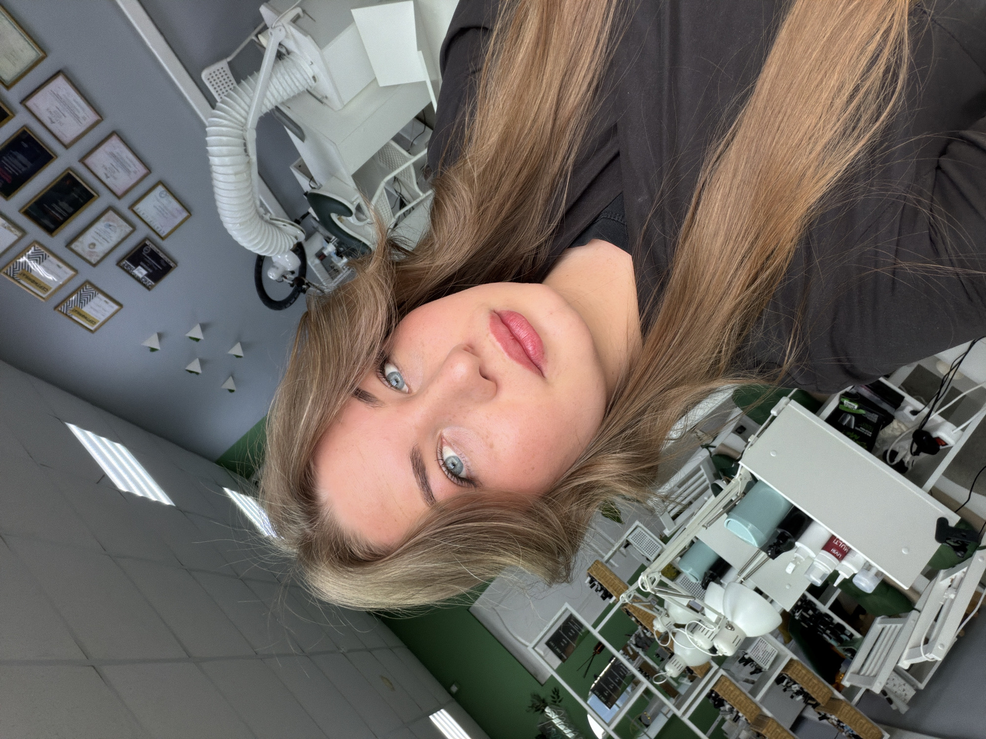 The image is a rotated selfie featuring a young woman looking directly at the camera with a calm, pleasant expression. She has long, straight, light brown hair, striking blue eyes, and wears light makeup, including pink lipstick and well-defined eyebrows. She is dressed in a dark, possibly black, long-sleeved top.
The setting is an indoor professional space, strongly suggesting a beauty salon or aesthetician's office in Chaikovsky, Russia. The background features a grey wall adorned with multiple framed certificates or diplomas, arranged in an irregular grid pattern. Below these, several small, white, triangular decorative elements are visible.
The room is equipped with modern white furniture and professional equipment. A prominent white machine with a flexible, ribbed hose, possibly a facial steamer or magnifying lamp, is visible. White tables or trolleys hold various supplies; one bottle has a red label with the Cyrillic text "УВЛАЖН" (UVLAZHN - Moisturizing/Hydrating) and "MULTI" clearly visible. Green wall sections add an accent to the decor. The ceiling features bright fluorescent lighting, illuminating the clean and professional environment.