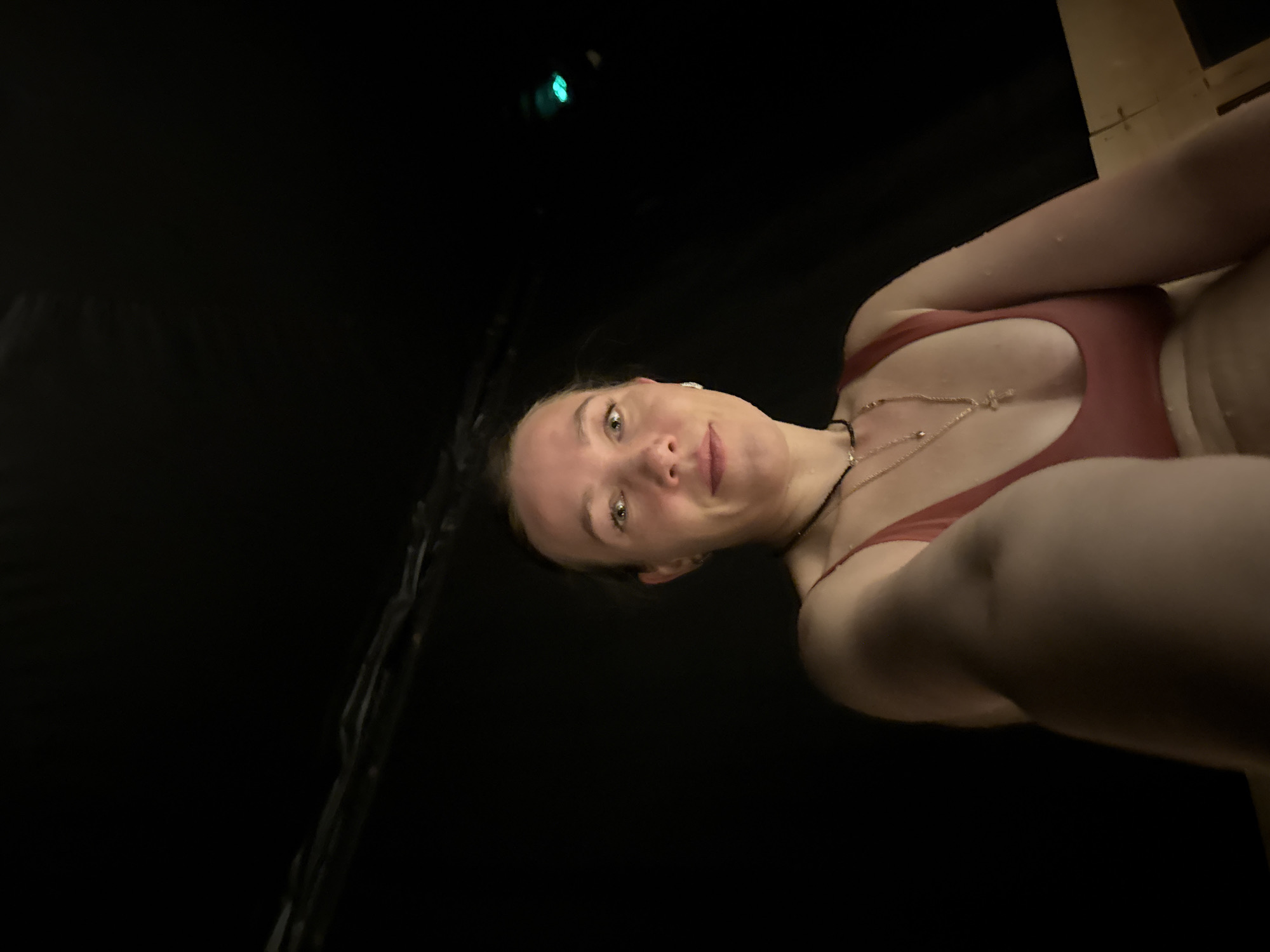 A low-angle selfie taken by a woman with fair skin and light brown hair pulled back, wearing a reddish-brown swimsuit and two necklaces, one with a cross pendant. The background is mostly black, with a dark green light visible at the top. The woman appears to be in a dark, possibly enclosed space, potentially a sauna due to the presence of what could be wooden planks and water droplets. The scene could be located in Beppu, Japan, given it is known for its onsen (hot springs) and associated facilities, such as saunas.