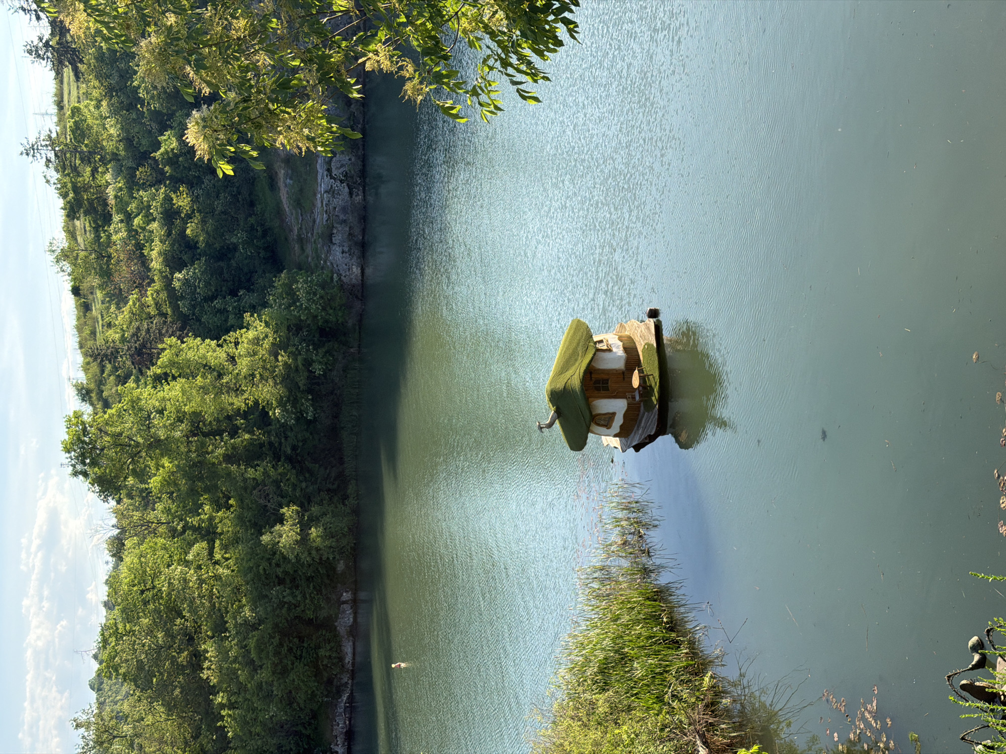 The image shows a small decorative house floating on a lake. The house has a green, thatched-looking roof and appears to be constructed of natural materials. It features a small window and what seems to be a miniature seating area. The lake is surrounded by lush green vegetation, including trees, bushes, and tall grass. The scene is bathed in natural light, suggesting it is daytime. The location is Lukovit, Bulgaria. There is a small white bird swimming in the lake, near the left side.