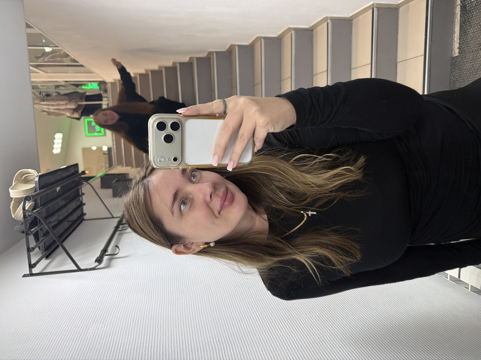 The image depicts a mirror selfie taken indoors. A woman in the foreground holds a gold-framed smartphone with a white back to capture her reflection. She has light brown hair with highlights, blue eyes, light pink lipstick, a gold necklace with a cross pendant, and a gold hoop earring with a pearl drop. Her right hand, holding the phone, displays light pink French-tipped nails and a thin dark green ring on the ring finger. She wears a black long-sleeved dress or top.
In the background reflection, a second individual with long, lighter brown hair and a dark top is visible on a light-colored staircase with gray risers. This individual is smiling and has her right arm extended horizontally. The environment includes a white textured wall, an antique-style dark metal coat rack with hooks and a shelf holding light-colored items, and bright green rectangular lights visible further back in the reflection. The specific location within the city of Chaikovsky, Russia, is not discernible from the provided image.