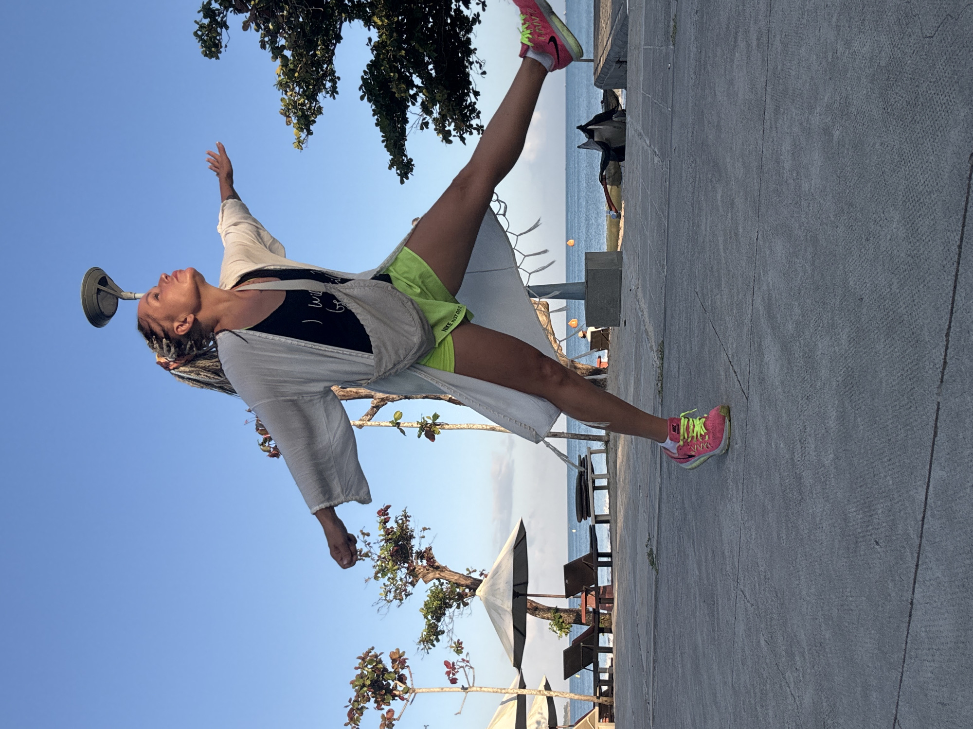 A woman with blonde braids and a white duster is captured mid-jump on a paved walkway near Kuta Selatan, Indonesia. She is wearing a black tank top with white text and neon yellow athletic shorts. Her arms are outstretched, and her right leg is extended forward, while her left leg is bent behind her. Bright pink and yellow athletic shoes adorn her feet. In the background, beach umbrellas and lounge chairs are visible on a sandy beach, with the ocean and a clear blue sky completing the scene. The lighting suggests it is daytime.