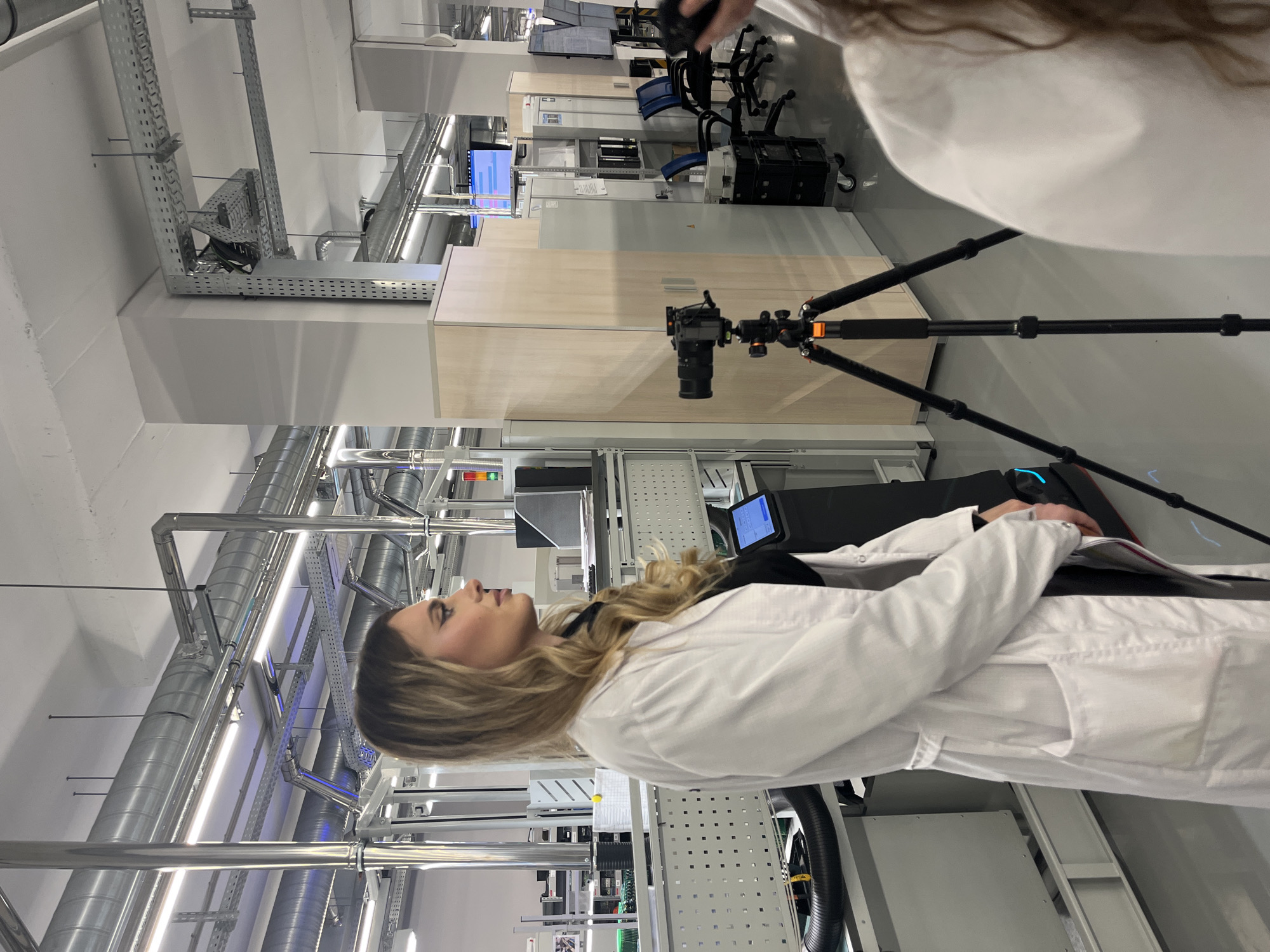 An indoor scene, likely in a modern industrial or laboratory facility located in Zelenodol'sk, Russia, is depicted. A woman with blonde hair, wearing a white lab coat, stands prominently in the left foreground, looking intently to her left (viewer's right) with a neutral and focused expression. Part of another person, also in a white lab coat, is visible in the upper right background.
The setting features a high ceiling with extensive exposed utilities, including large silver pipes, ventilation ducts, and metal cable trays, characteristic of a functional, high-tech environment. Bright artificial lighting illuminates the space. In the mid-ground, a black camera is mounted on a tripod, suggesting filming or photography is in progress, potentially documenting the facility or an ongoing activity.
The background reveals various pieces of industrial or laboratory equipment, including machinery or workstations with monitors displaying light blue interfaces, along with office furniture such as black chairs and light wooden cabinets. The overall atmosphere is professional, clean, and organized, typical of a research lab, a factory floor, or an educational facility focused on technical operations. The visible activity suggests a formal observation, demonstration, or a recording session within this specialized environment. No specific text is legible on any surfaces or screens.