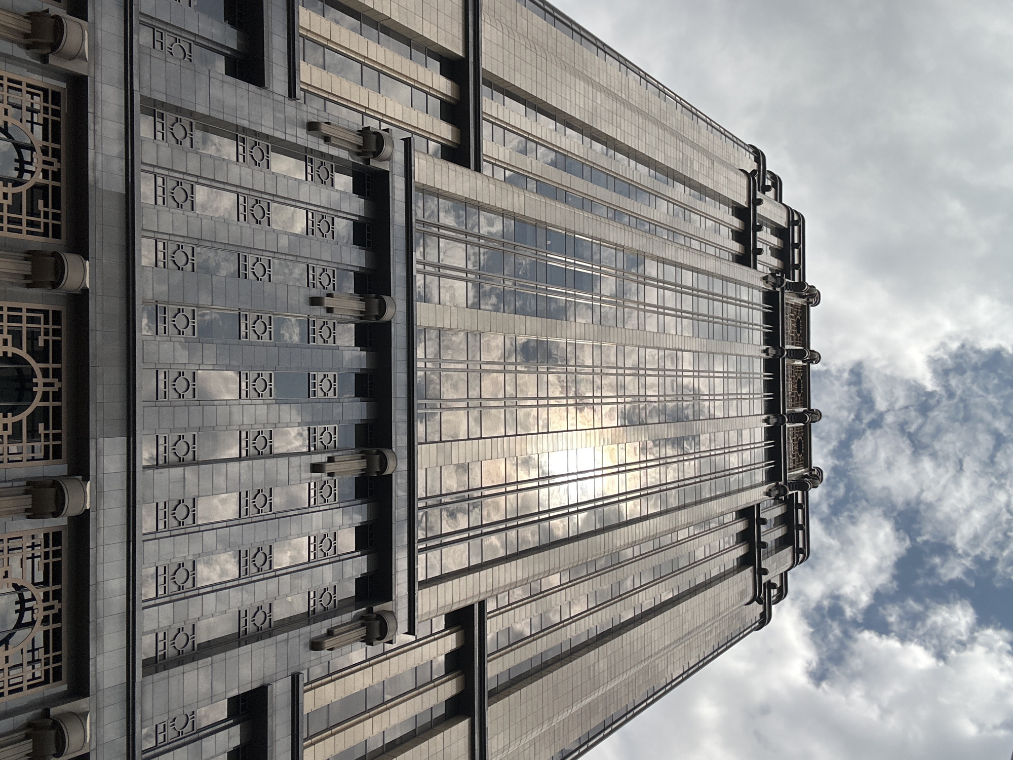 A low-angle shot captures the facade of a tall, art deco style skyscraper in Singapore. The building's exterior is a mix of polished stone and reflective glass, with intricate geometric patterns adorning its lower levels. The glass panels reflect the cloudy sky, with patches of sunlight breaking through. The skyscraper's upper section features tiered setbacks and decorative elements, typical of art deco architecture. The overall impression is one of grandeur and a bygone era of architectural design.