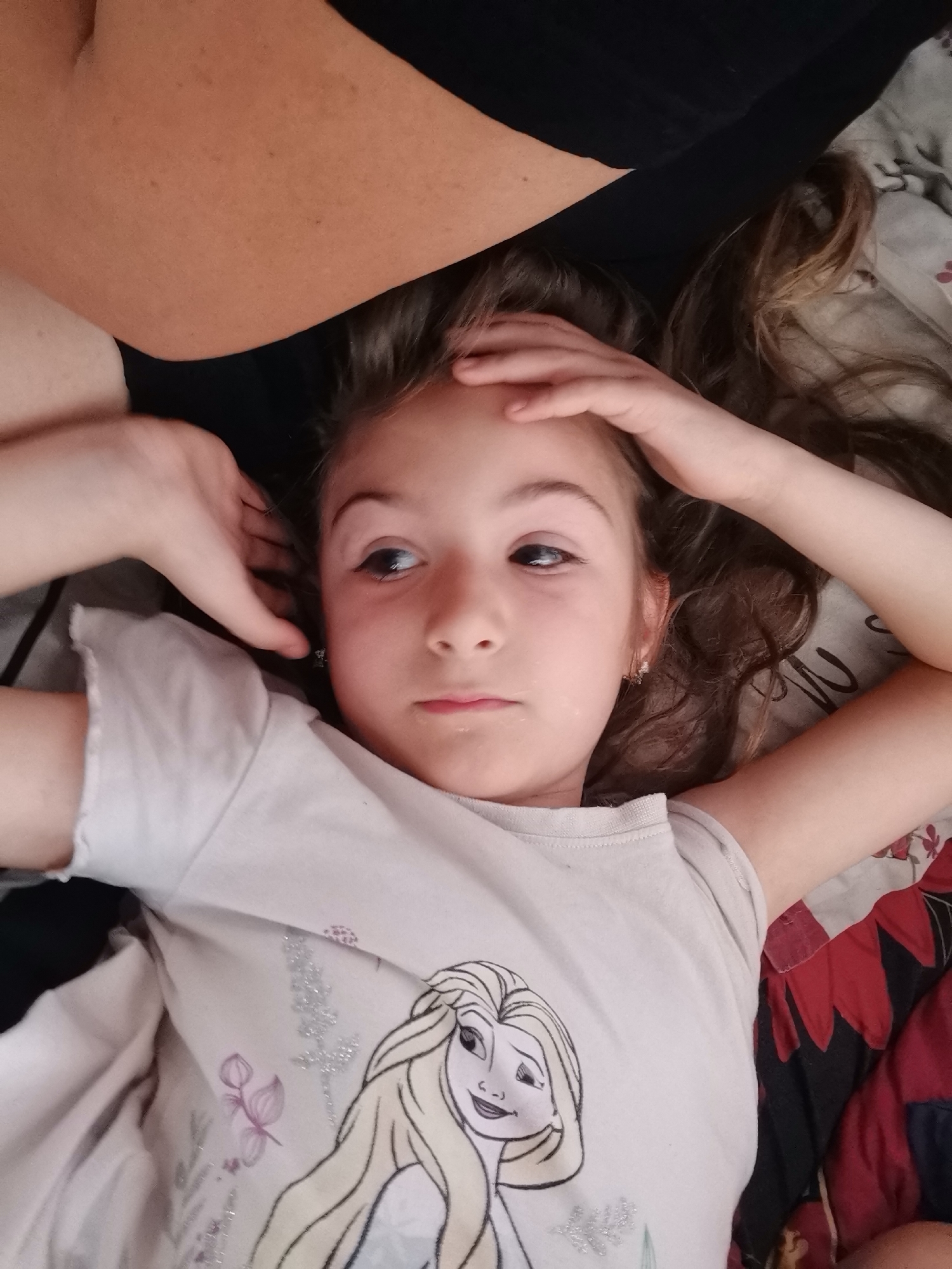 A close-up, high-angle shot depicts a young girl, likely between 5 and 10 years old, lying down indoors, presumably on a bed or couch. She has light brown hair and is wearing a light-colored t-shirt adorned with an illustration of Elsa from Disney's Frozen, along with glittery accents and subtle floral designs. Her hands are positioned behind or near her head, suggesting a relaxed or contemplative posture. She gazes towards the upper right of the frame with a somewhat neutral, slightly wide-eyed expression. A small earring is visible in her right ear.
In the upper left portion of the image, part of an adult's arm or shoulder, dressed in a black garment, is visible, resting close to the girl's head. The bedding or fabric beneath her features a distinct red and black floral or leaf pattern on the right side, transitioning to a lighter, more subdued pattern towards the left. The indoor lighting appears soft and even. No explicit text is visible in the image. The precise location is unknown.