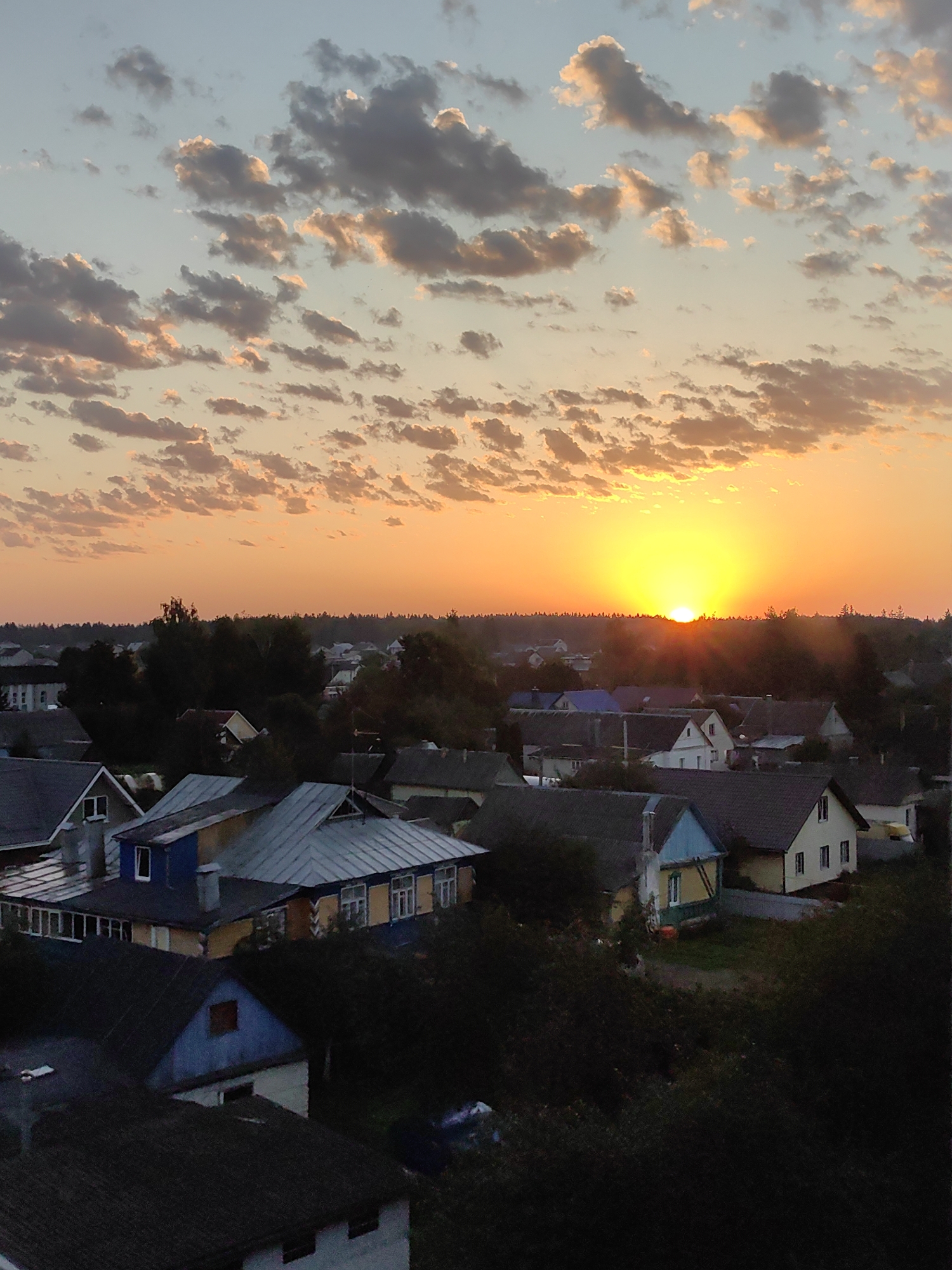 A high-angle, full-frame, vertical, medium shot captures a sunset over a village, likely in Fanipal, Belarus, as indicated by similar imagery. The sky dominates the upper two-thirds of the frame, with the sun, a bright yellow orb, positioned just above the horizon line of trees in the mid-ground. Rays of light emanate from the sun, casting an orange and yellow glow across the sky. Wispy, scattered clouds are illuminated by the setting sun, appearing in shades of gray, white, and pink. The lower third of the image depicts a village with numerous houses. The houses are of various sizes and architectural styles, with pitched roofs of gray metal or dark shingles. Some houses have brightly colored walls, including yellow, blue, and white. Trees and dense foliage are interspersed among the houses, partially obscuring some of the structures. The overall scene conveys a peaceful, rural atmosphere at dusk. There are no people or specific actions depicted.